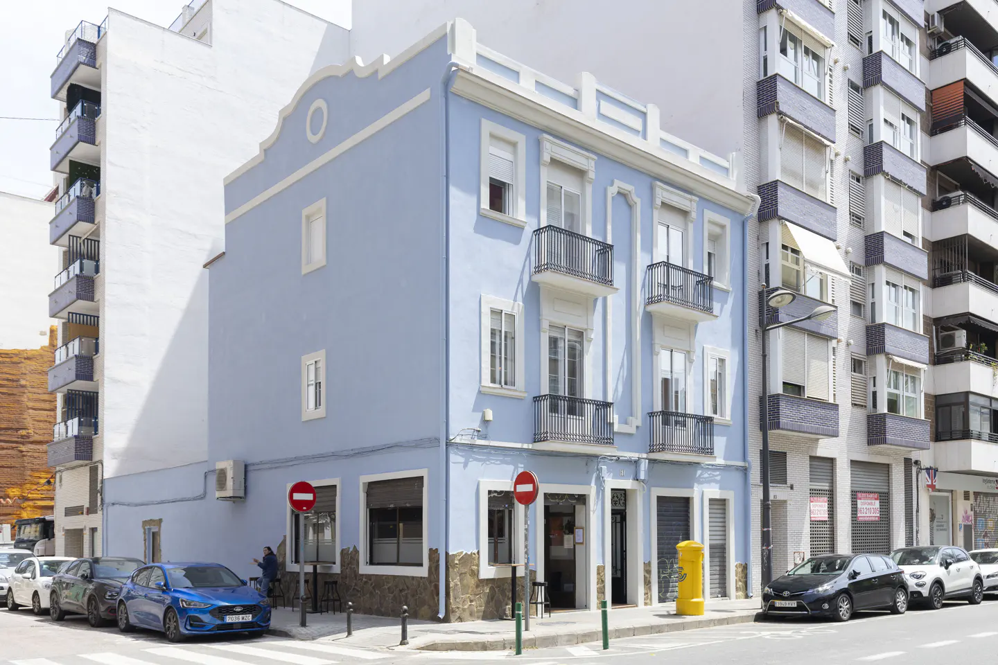A three-story light blue building with white trim and small balconies, located on a city street corner.