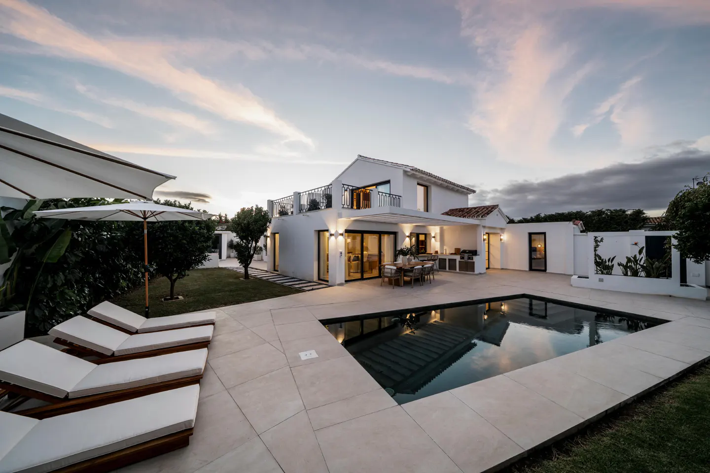 Modern white villa with a swimming pool, patio lounge chairs, and outdoor dining area at sunset.