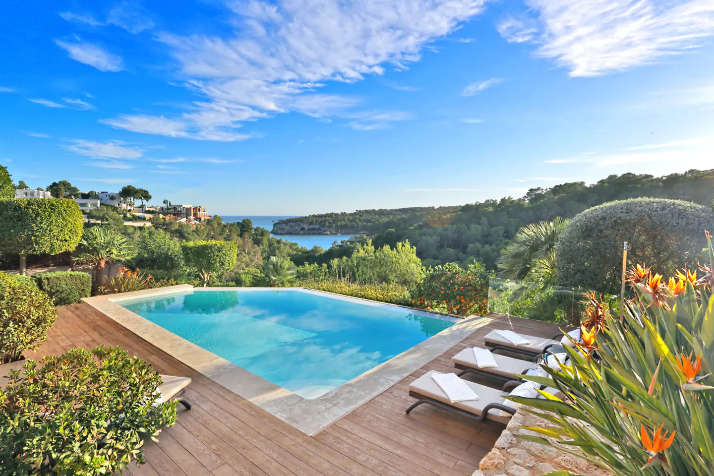 Outdoor pool with lounge chairs on a wood deck, overlooking a bay and green hills under a blue sky.