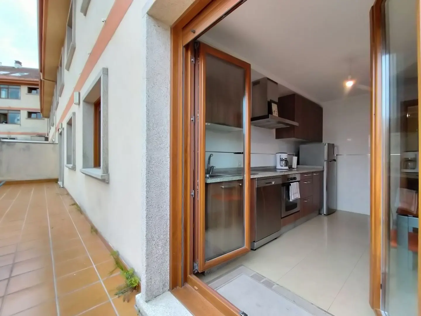 Open wood-framed glass doors lead from a tiled patio into a kitchen. The kitchen has stainless steel appliances and dark wood cabinets.