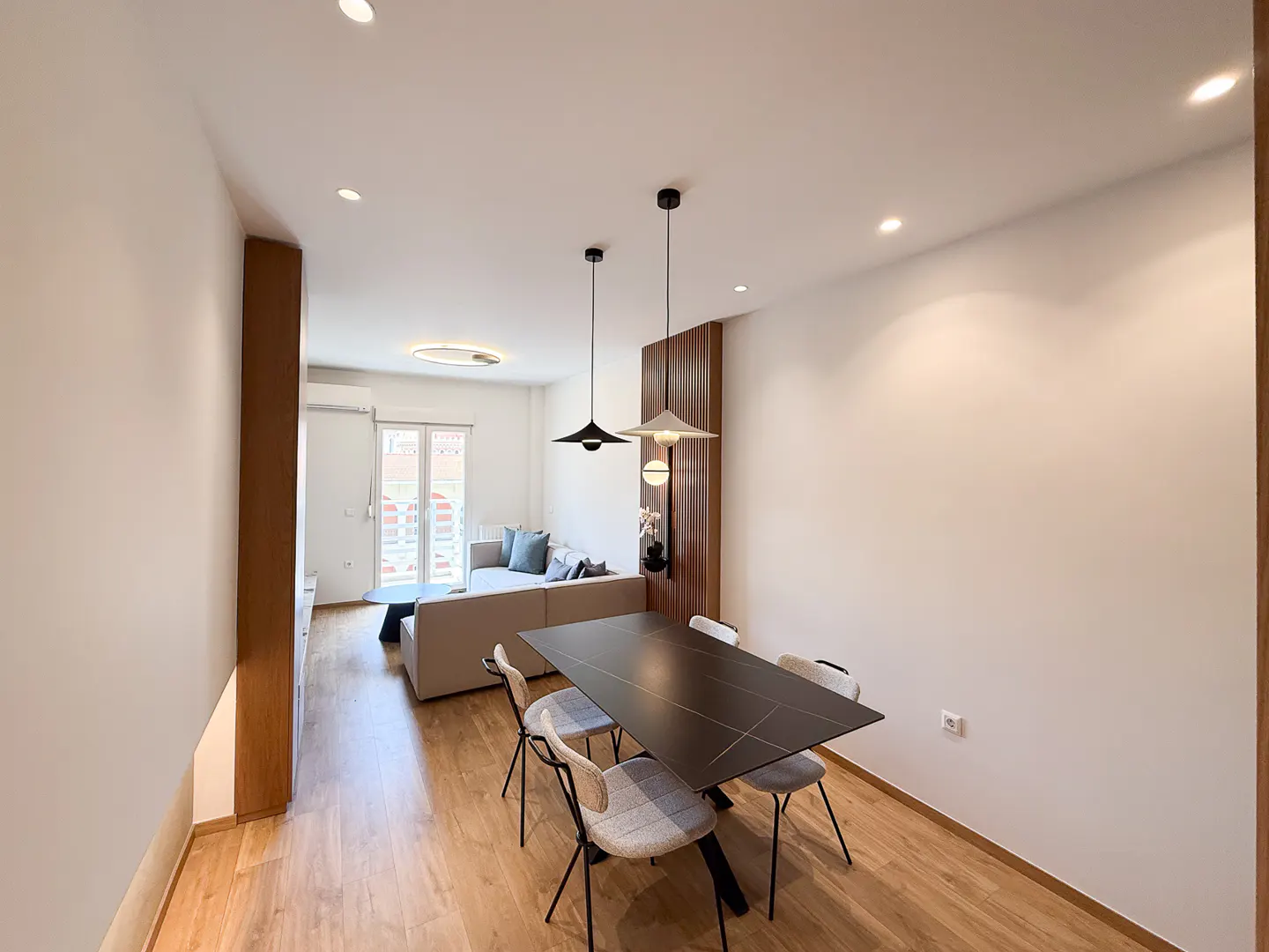 Bright, modern apartment with a black dining table and four chairs. A beige sofa is visible in the background near a window.