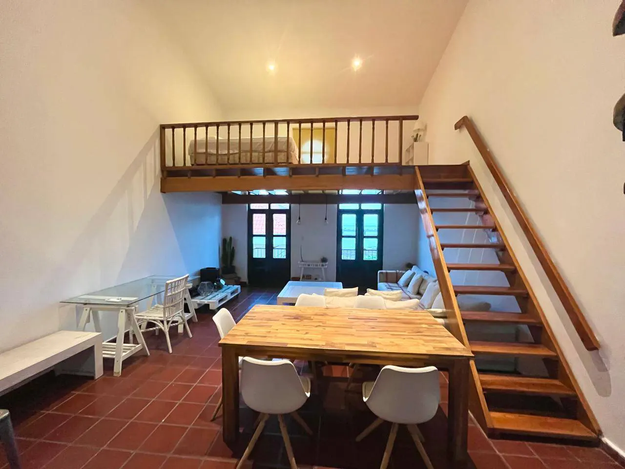 Open-plan living area with a wooden dining table, white chairs, terracotta floors, and a loft bedroom with wooden stairs.