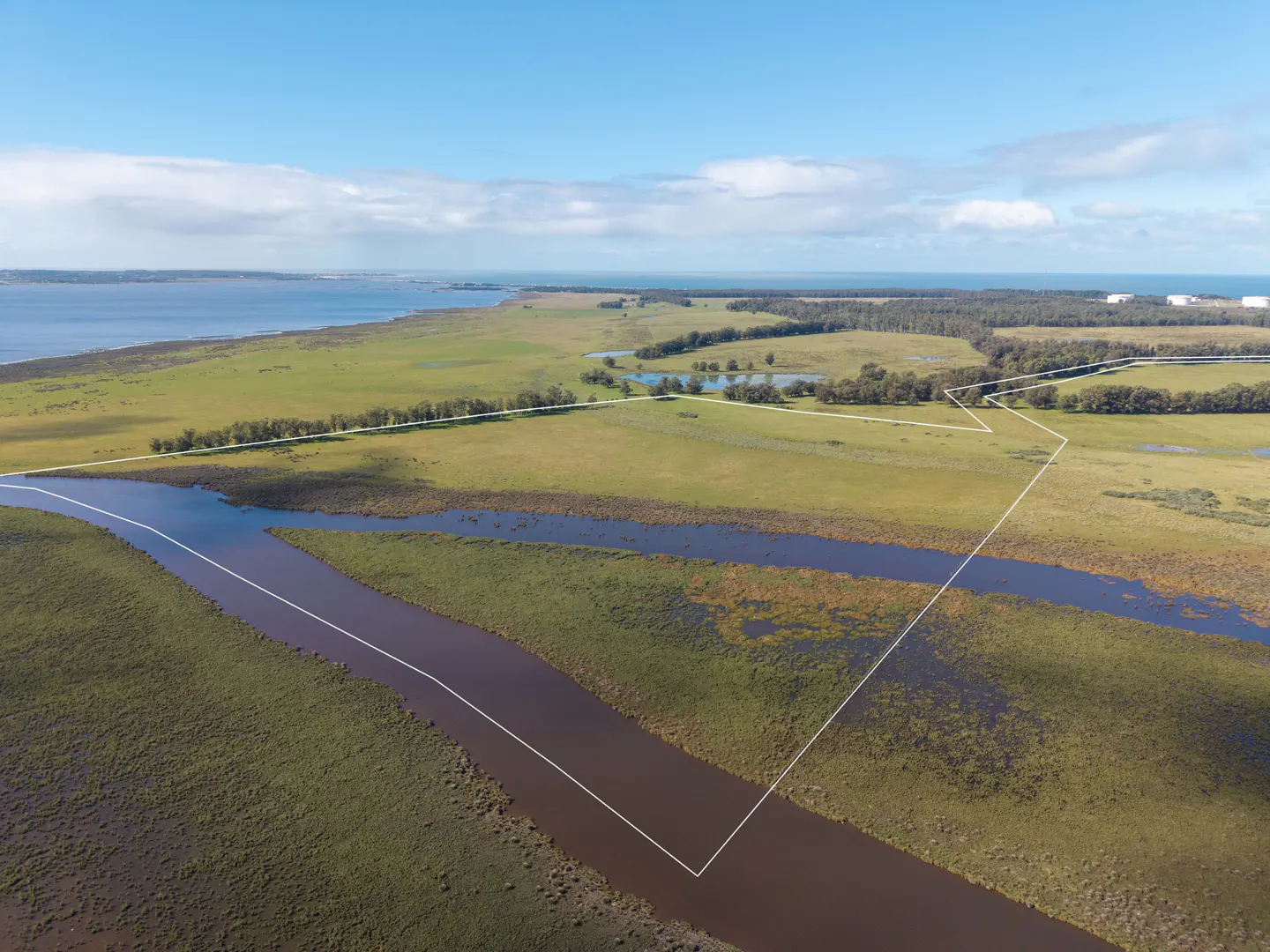 Aerial view of a large, green, undeveloped property with a river running through it, outlined in white. Blue sky and water in the background.