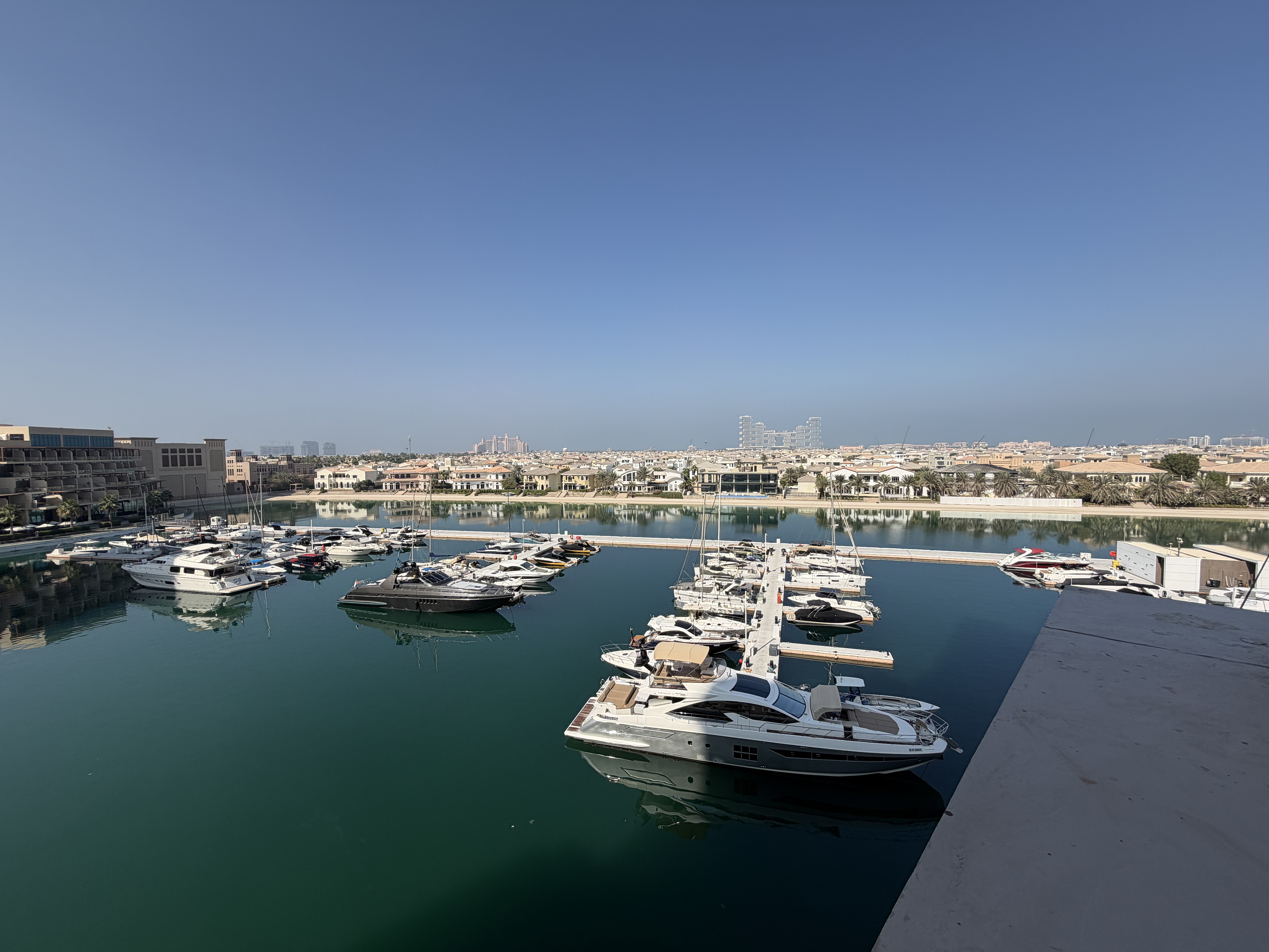 High-angle view of a calm marina with multiple luxury yachts docked in turquoise water under a clear blue sky.