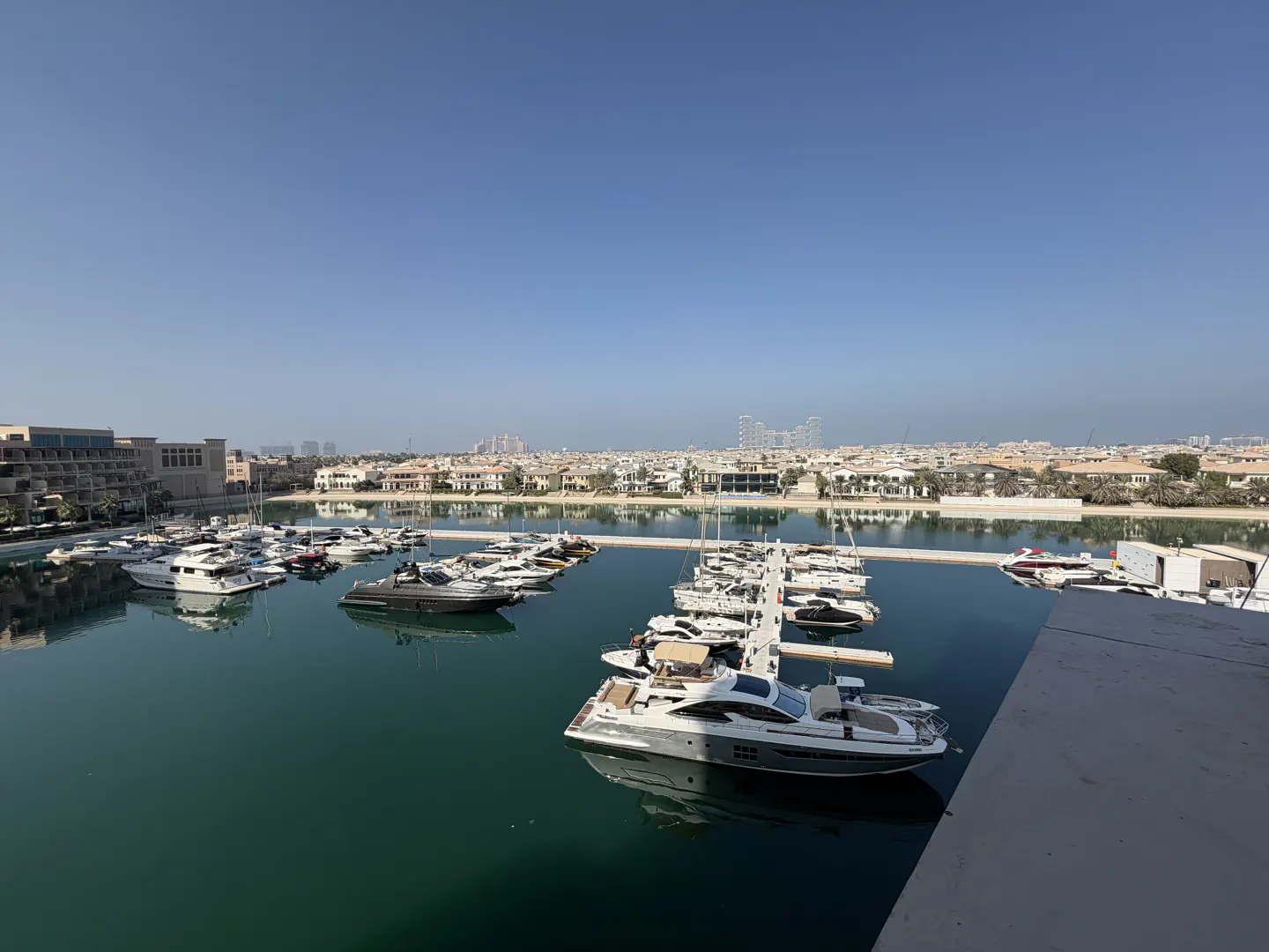 High-angle view of a calm marina with multiple luxury yachts docked in turquoise water under a clear blue sky.