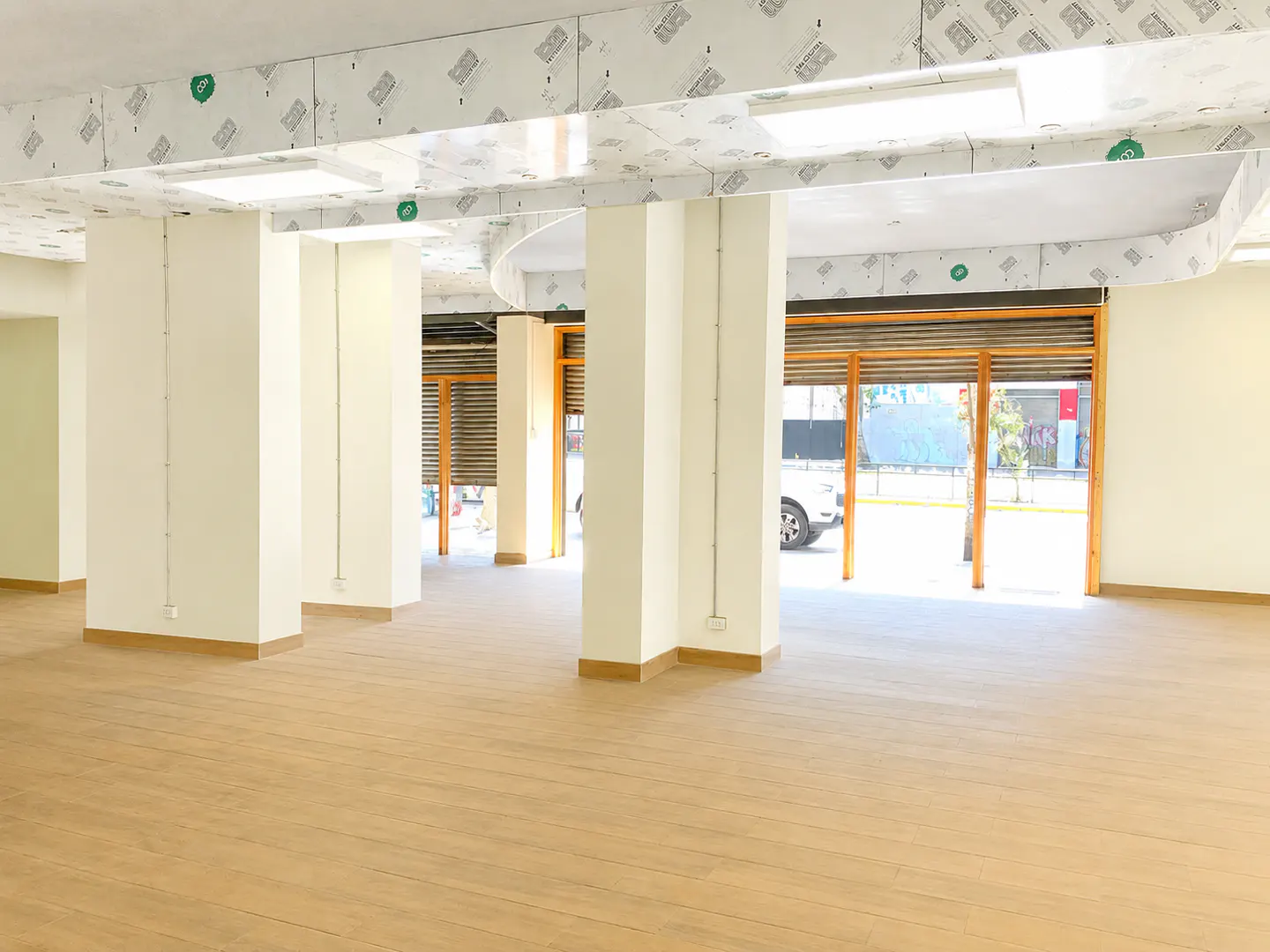 Empty commercial space with light wood floors, white support pillars, and large windows with metal roll-down shutters.