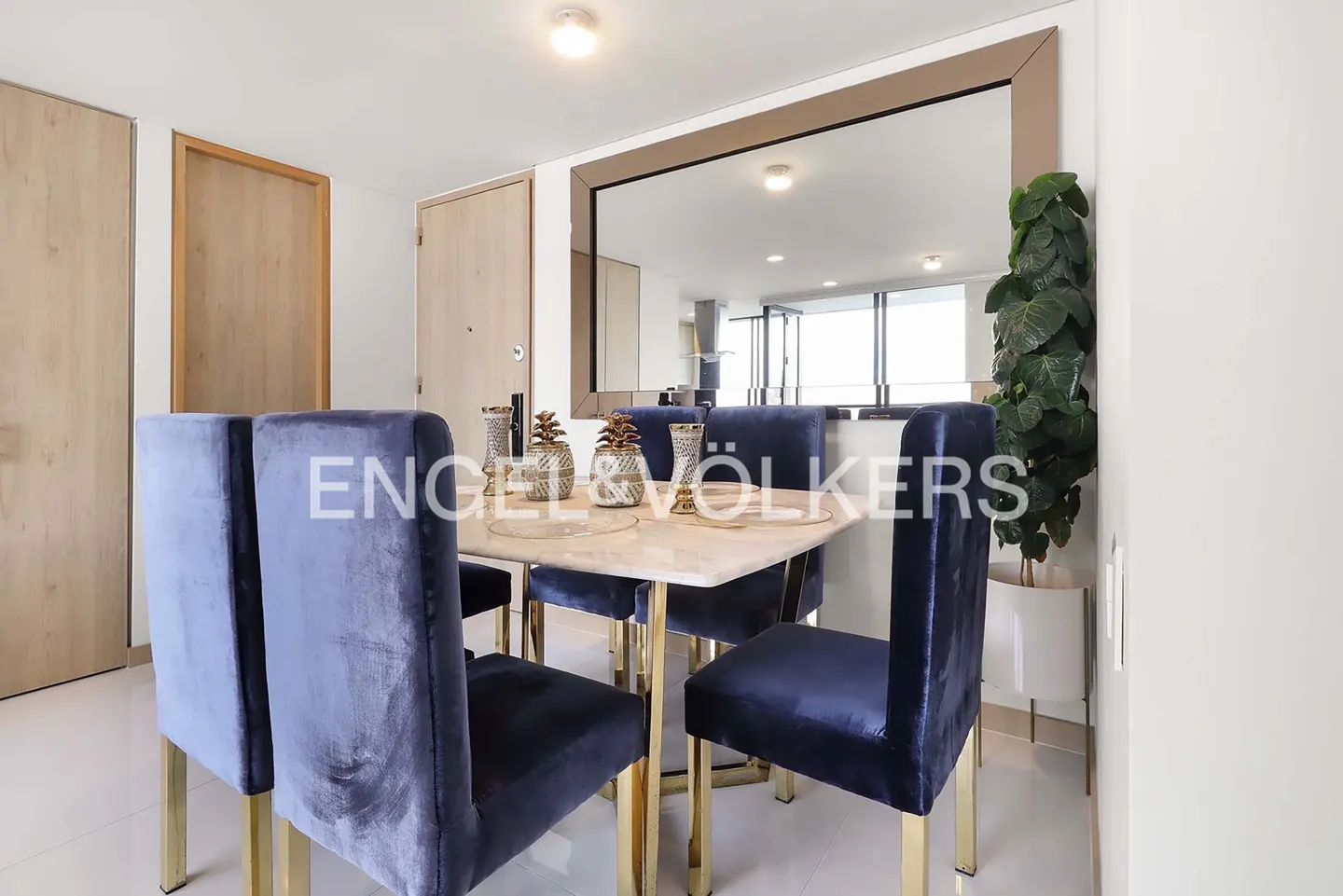 Dining area with a square marble table, four blue velvet chairs with gold legs, and a large mirror reflecting the room.