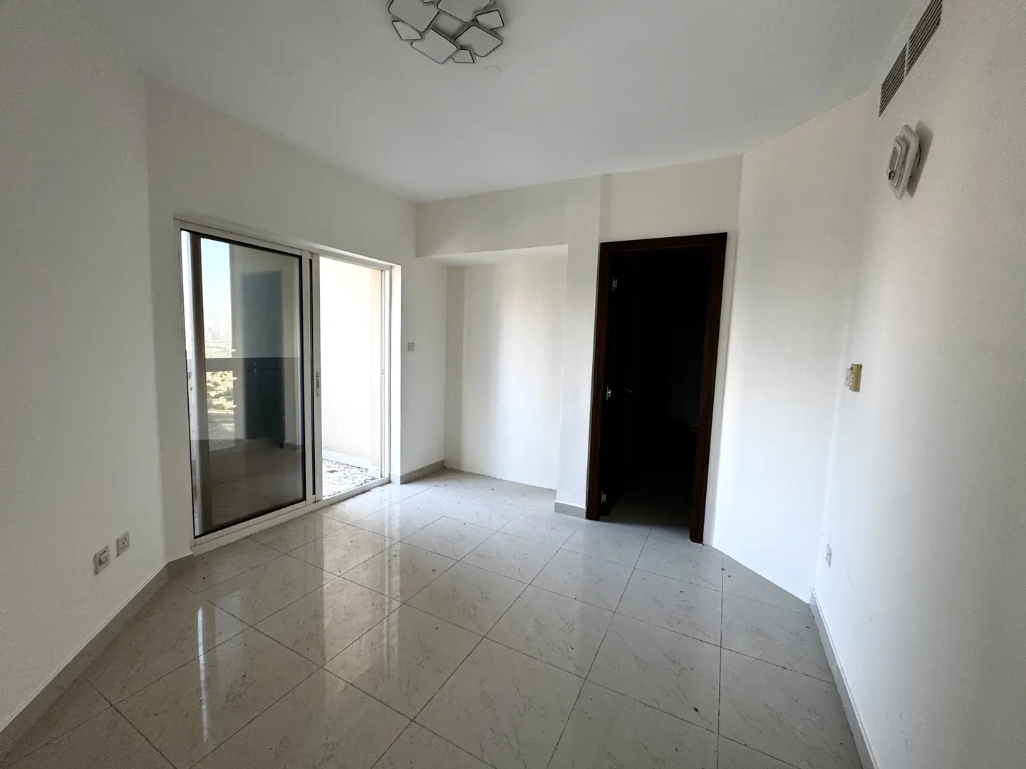 An empty room with white walls, light tile floor, and a sliding glass door to a balcony. A dark wood door is on the right.