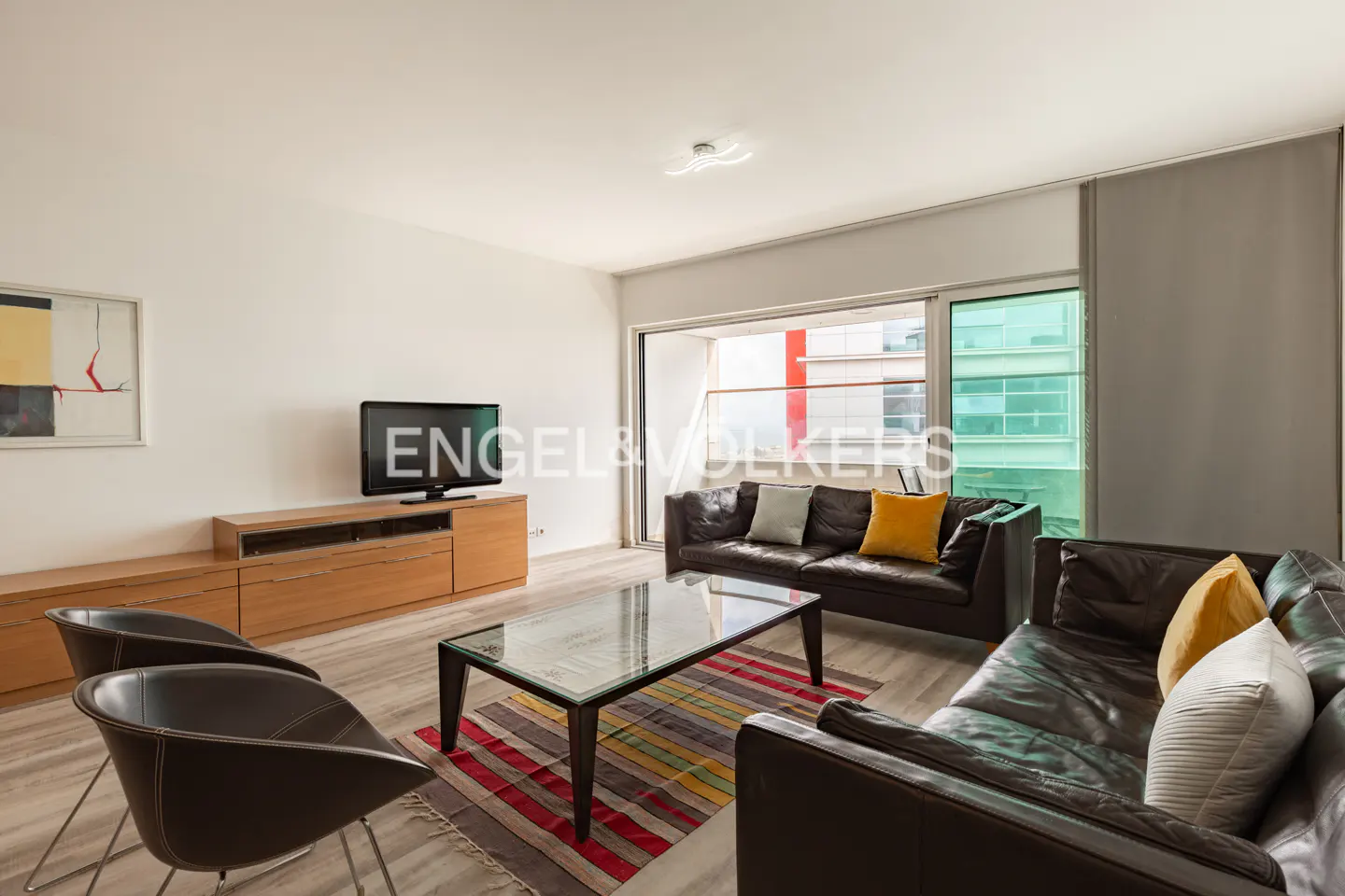 Living room with brown leather sofas, glass table, TV on wood cabinet, two chairs, and striped rug. Balcony view.