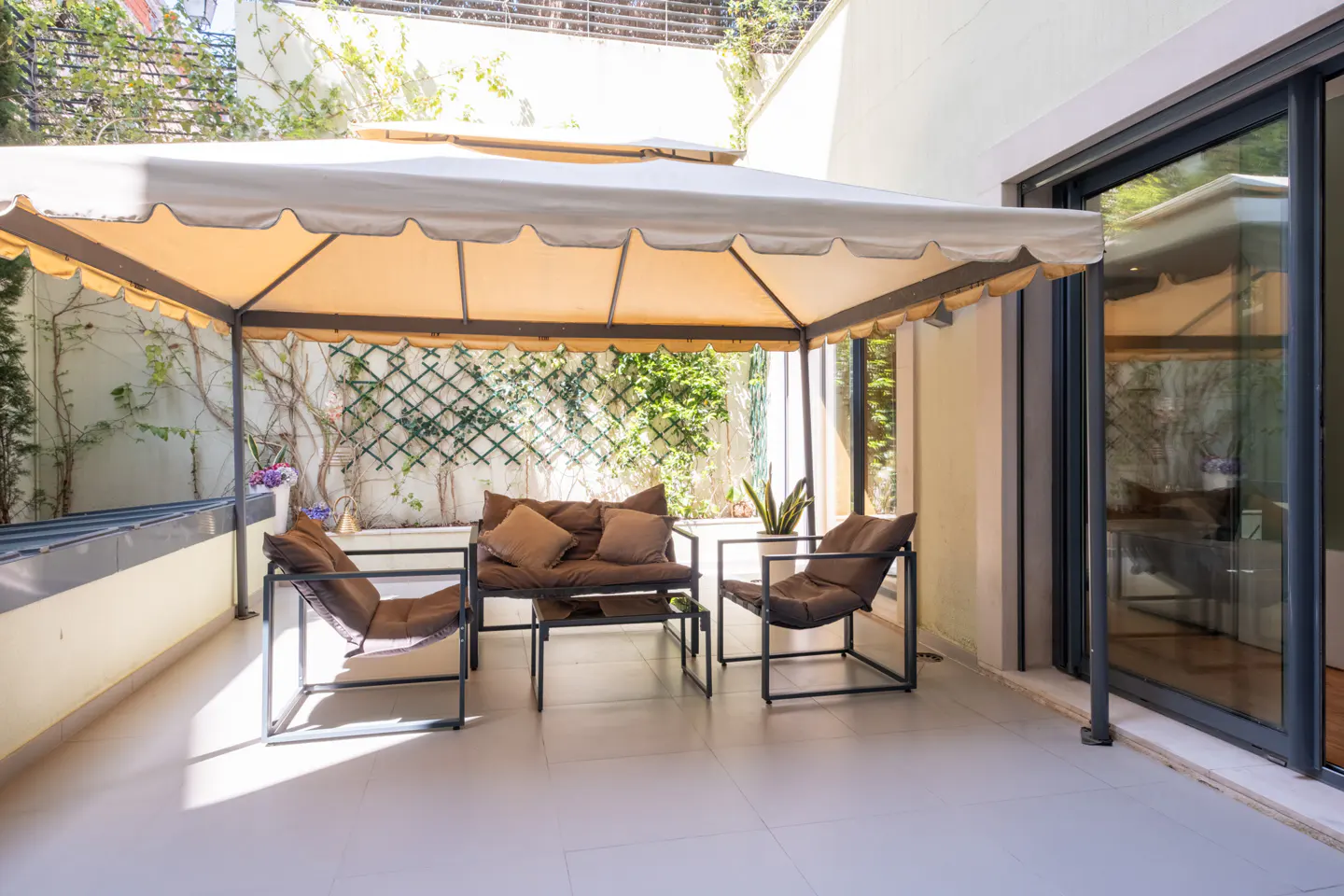 Outdoor patio with brown cushioned seating under a beige canopy. Green vines climb the white walls in the background. Sliding glass doors to the right.