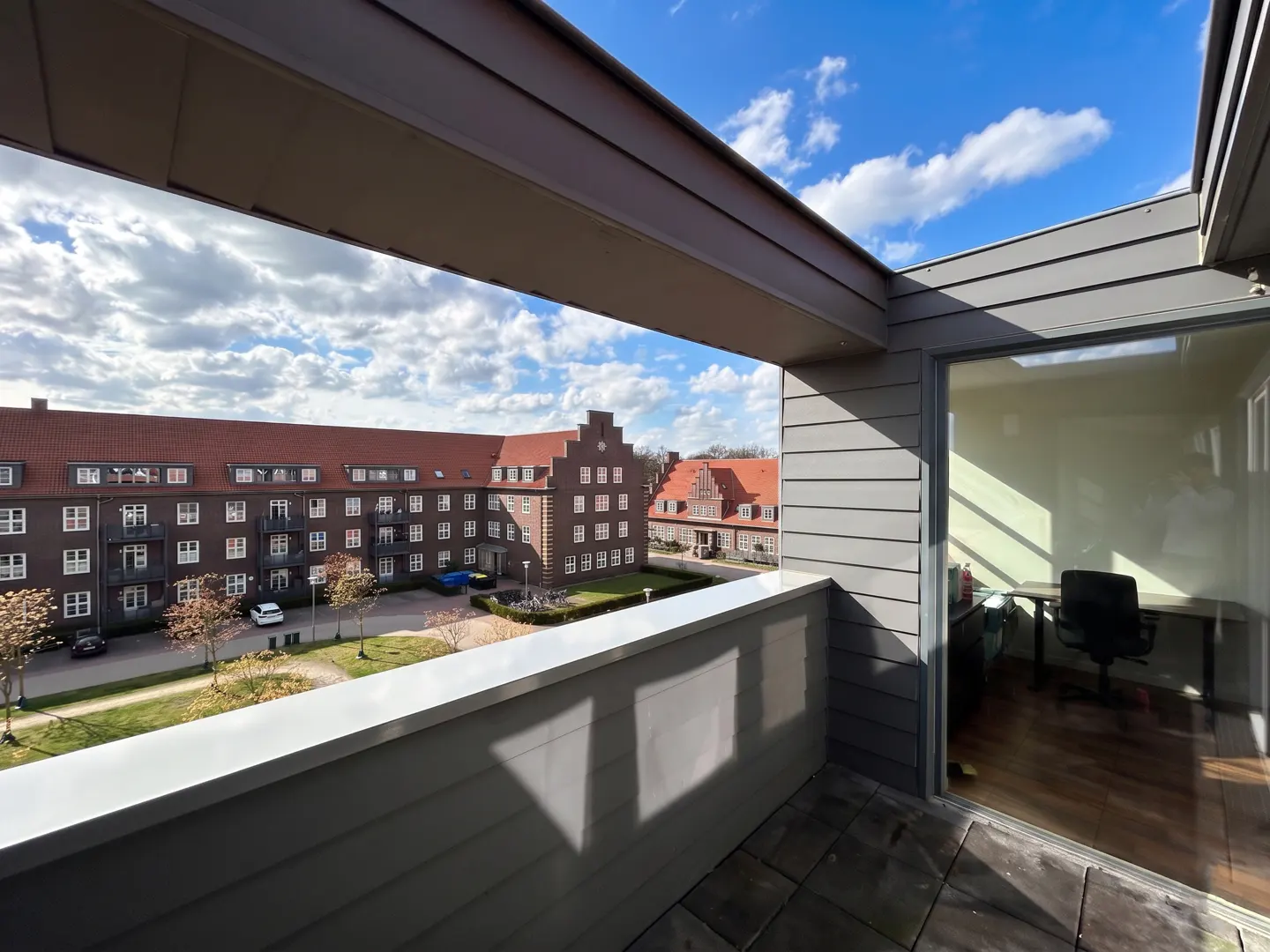 View from a gray balcony overlooking a brick apartment building with a red roof and a blue sky with white clouds. A home office is visible through a glass door.