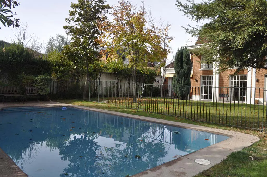 Backyard pool with a brick house in the background, surrounded by a black metal fence and trees.