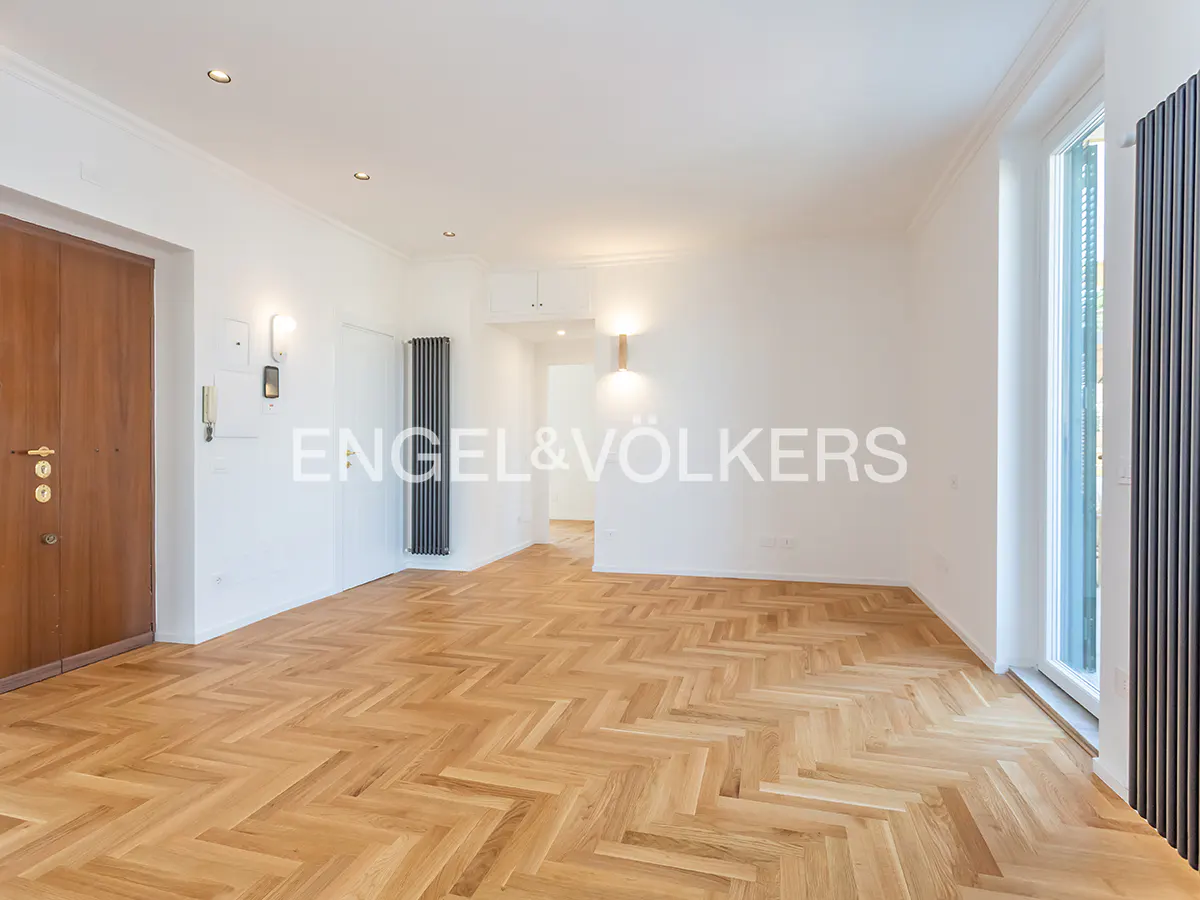 Bright, empty room with herringbone wood floors, white walls, and a brown door. A tall, dark radiator is near a window.