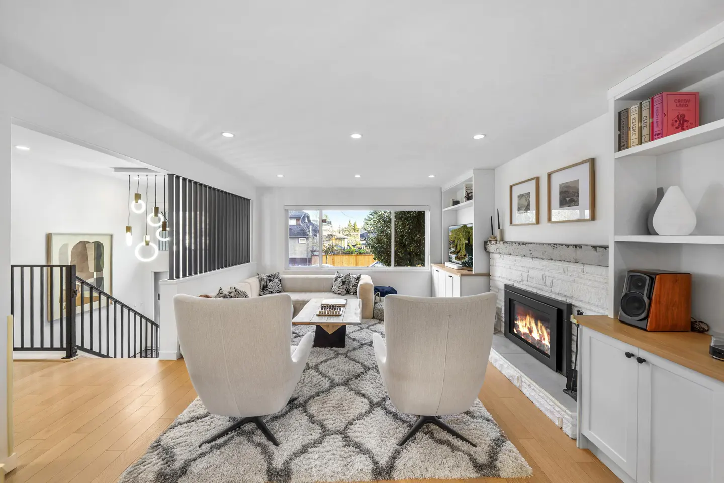 Bright living room with hardwood floors, white walls, and a patterned rug. Two white chairs face a fireplace with built-in shelves. A staircase is visible on the left.