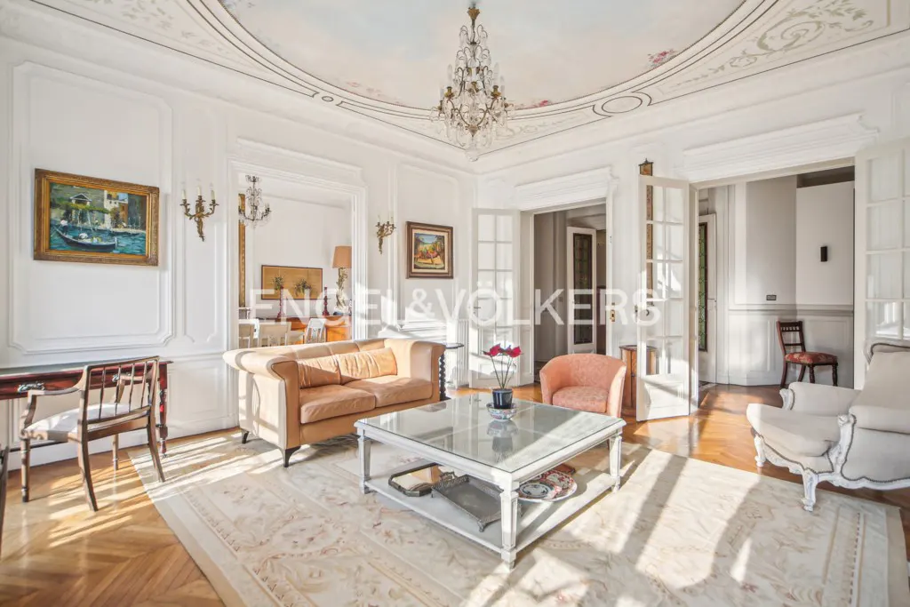 Elegant living room with white paneled walls, a crystal chandelier, tan sofa, glass coffee table, and parquet flooring.
