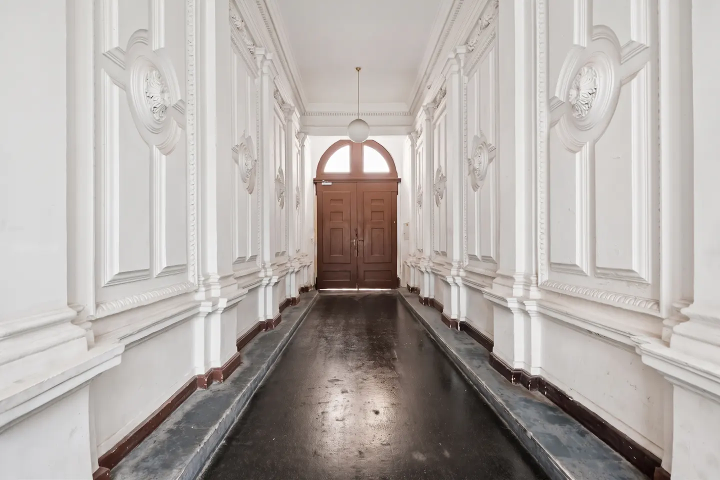 Hallway with white walls, ornate molding, and dark wood doors at the end. A globe light hangs from the ceiling.