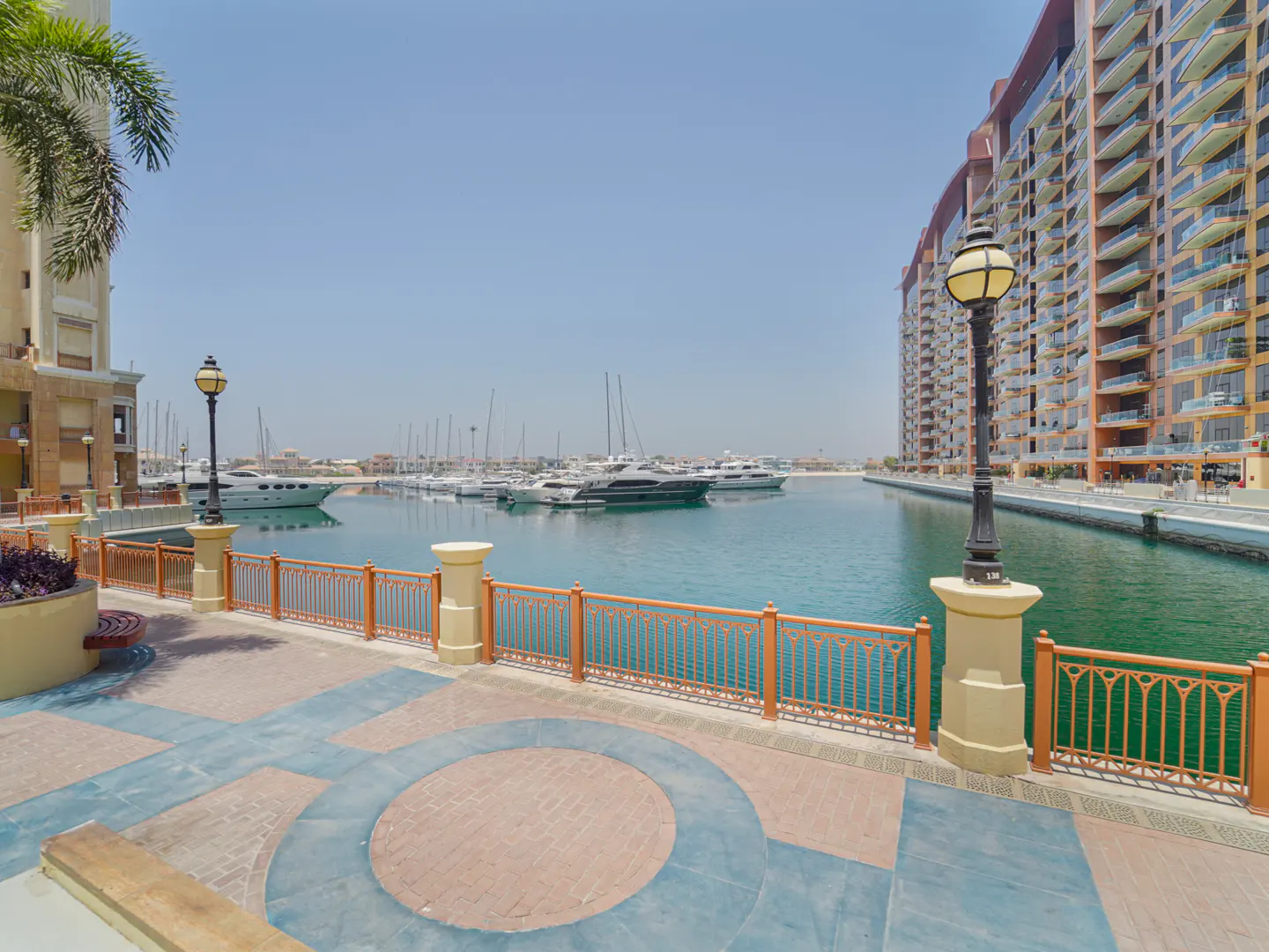 Waterfront view of a marina with yachts, buildings, and a patterned walkway with a decorative fence.