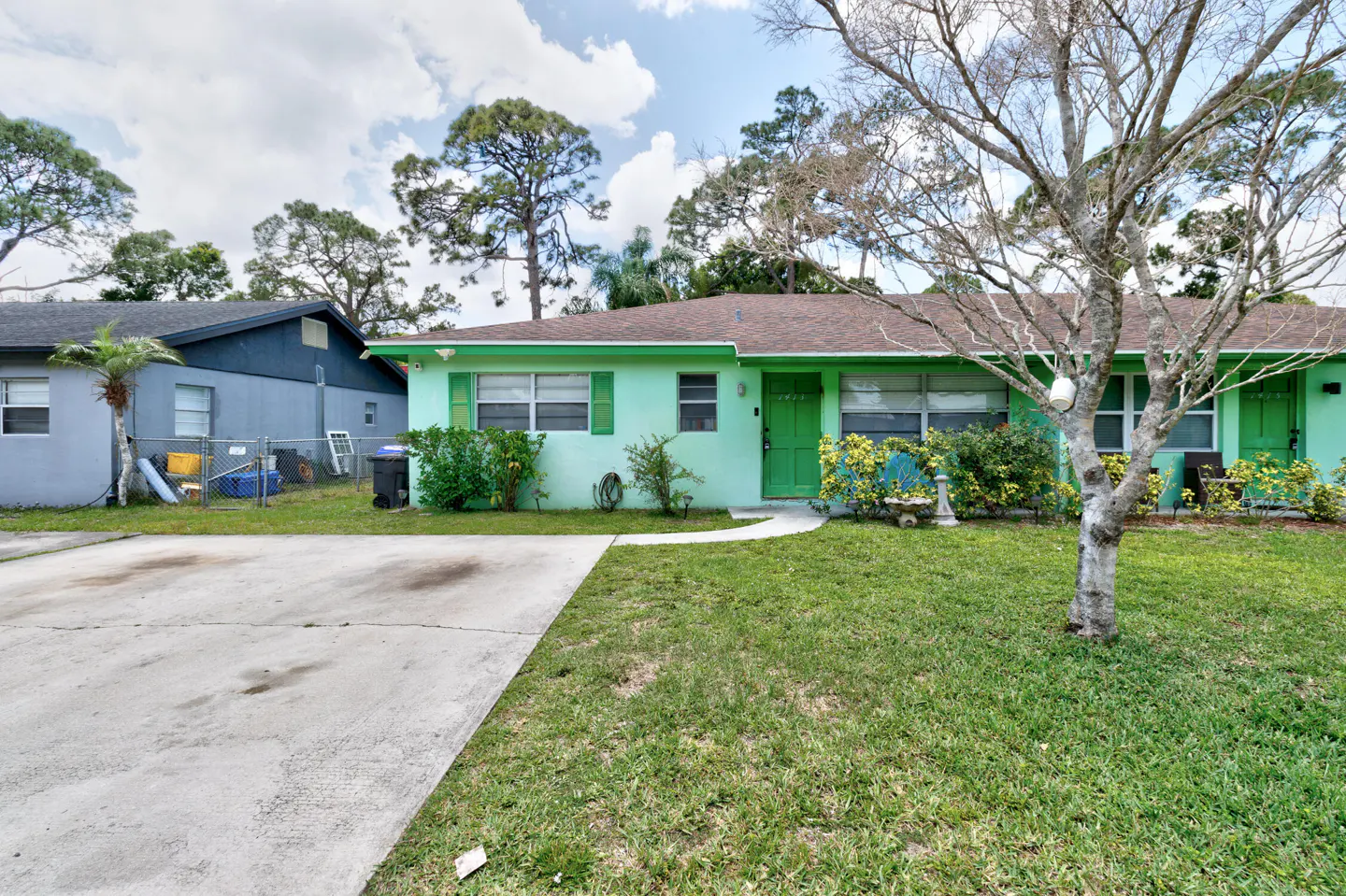A light green duplex with a brown roof, green shutters, and a concrete driveway. A bare tree stands in the front yard.