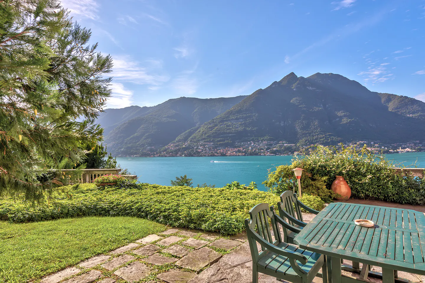 Outdoor patio with green table and chairs overlooking a blue lake and mountains on a sunny day.