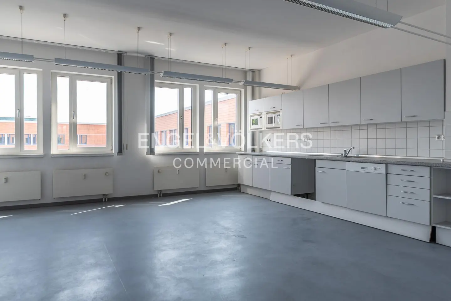 Bright, empty office kitchen with grey cabinets, white tiled backsplash, grey flooring, and large windows.