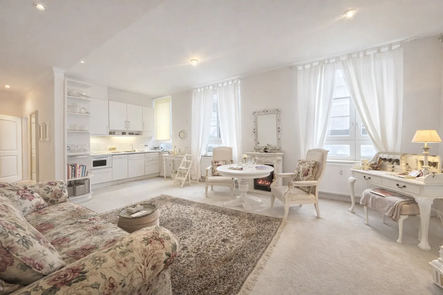 Bright, open-concept living space with white walls, kitchen, and floral sofa. A fireplace and desk add charm.