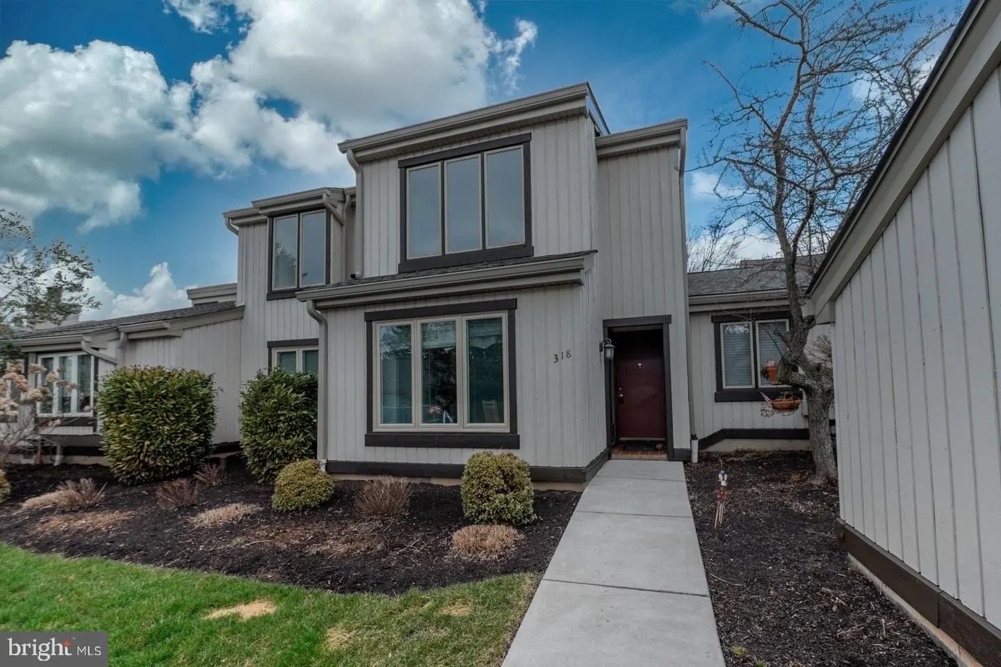 Exterior view of a two-story townhouse with light gray siding, dark brown trim, and a red front door. A concrete walkway leads to the entrance.