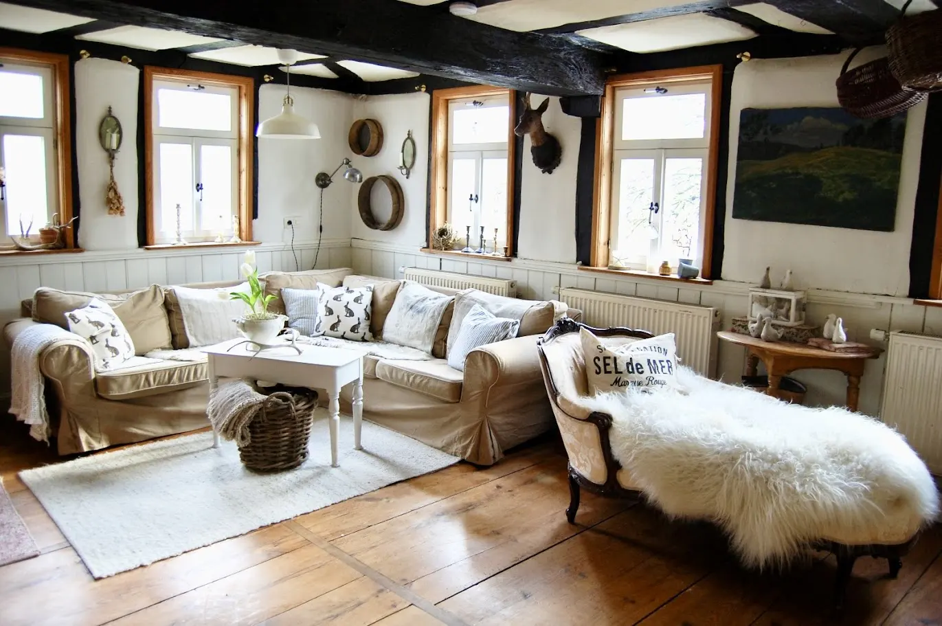 Cozy living room with beige sofa, white chaise lounge with fur throw, and wood floors. Black beams accent the white walls and window frames.