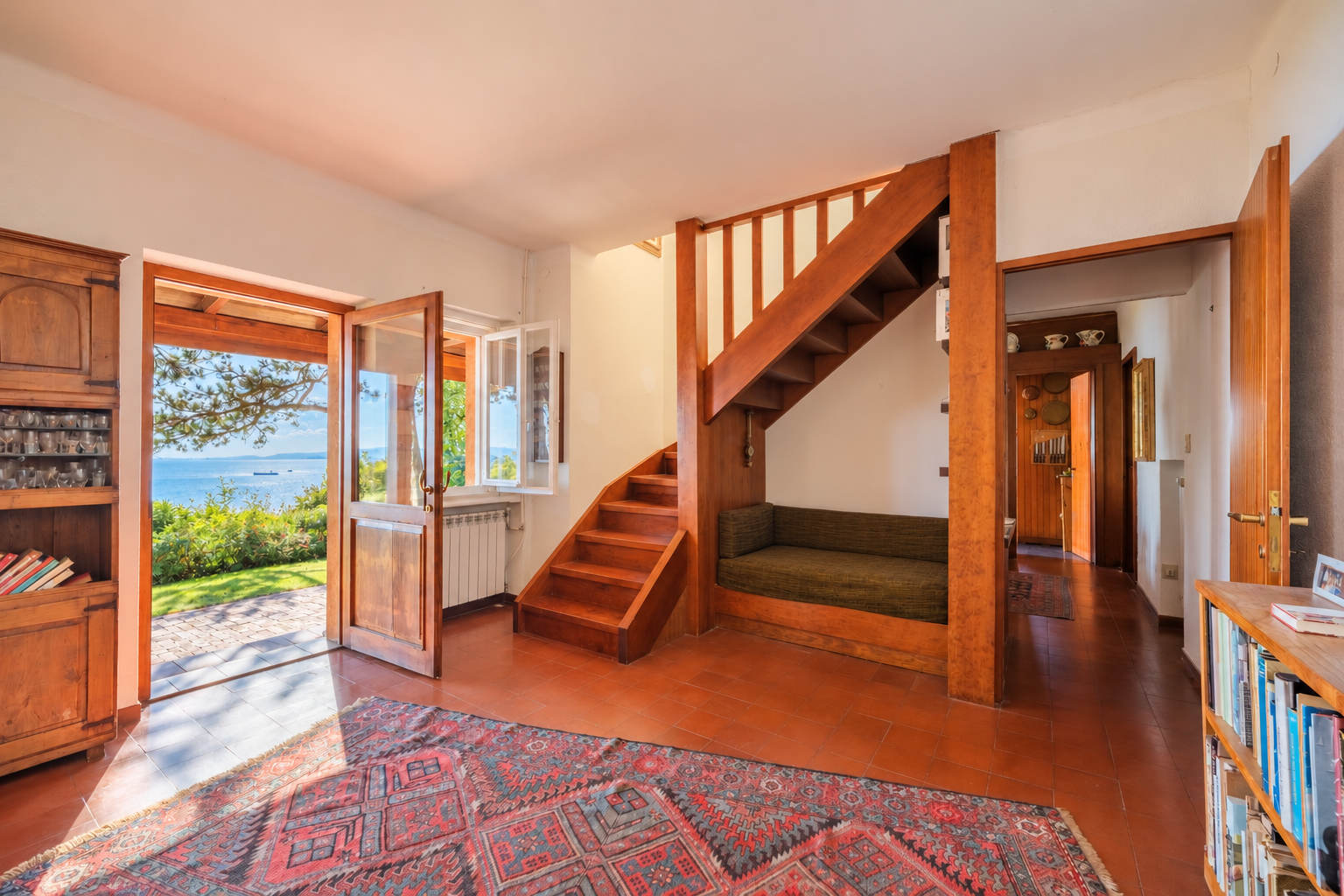 Living room with wooden staircase, built-in bench, patterned rug, and open door revealing a scenic ocean view. [AI-generated text]