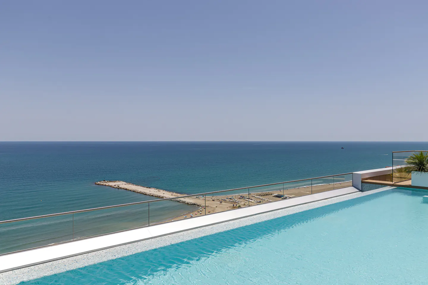 Rooftop pool with clear glass railing overlooks the ocean and beach on a sunny day.