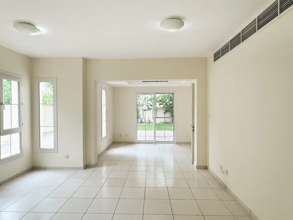 Empty, bright room with white tile floor, cream walls, and sliding glass doors to a green yard. Windows on the left.