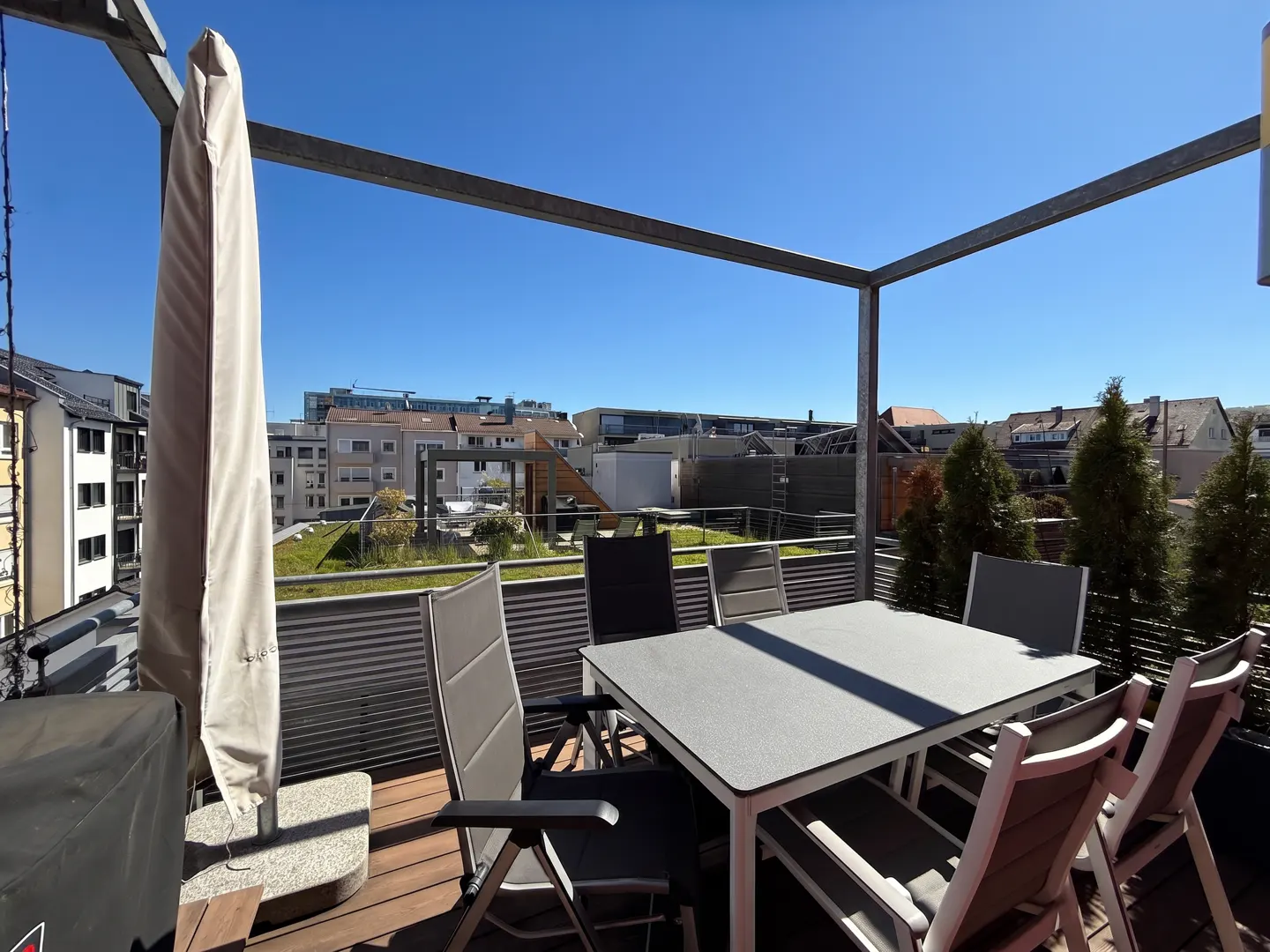 Outdoor patio with a table, chairs, and a closed umbrella. The patio has a wooden floor and a metal frame. Buildings are visible in the background.