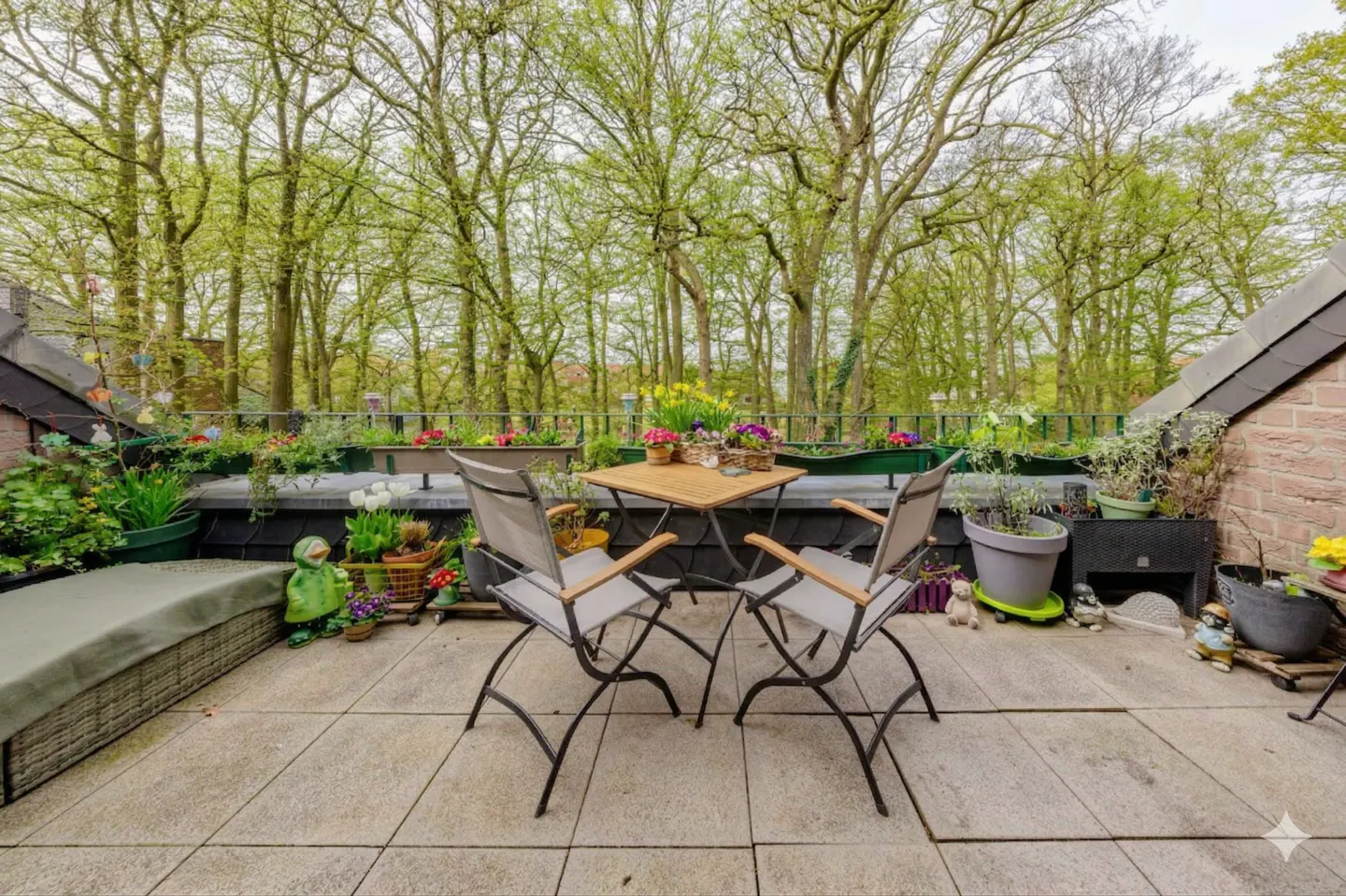 Rooftop patio with table, chairs, and potted plants. Trees are visible in the background.