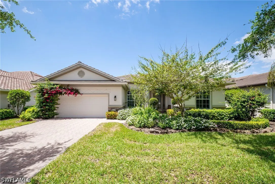 Beige single-story house with a white garage door, green lawn, and lush landscaping under a blue sky.