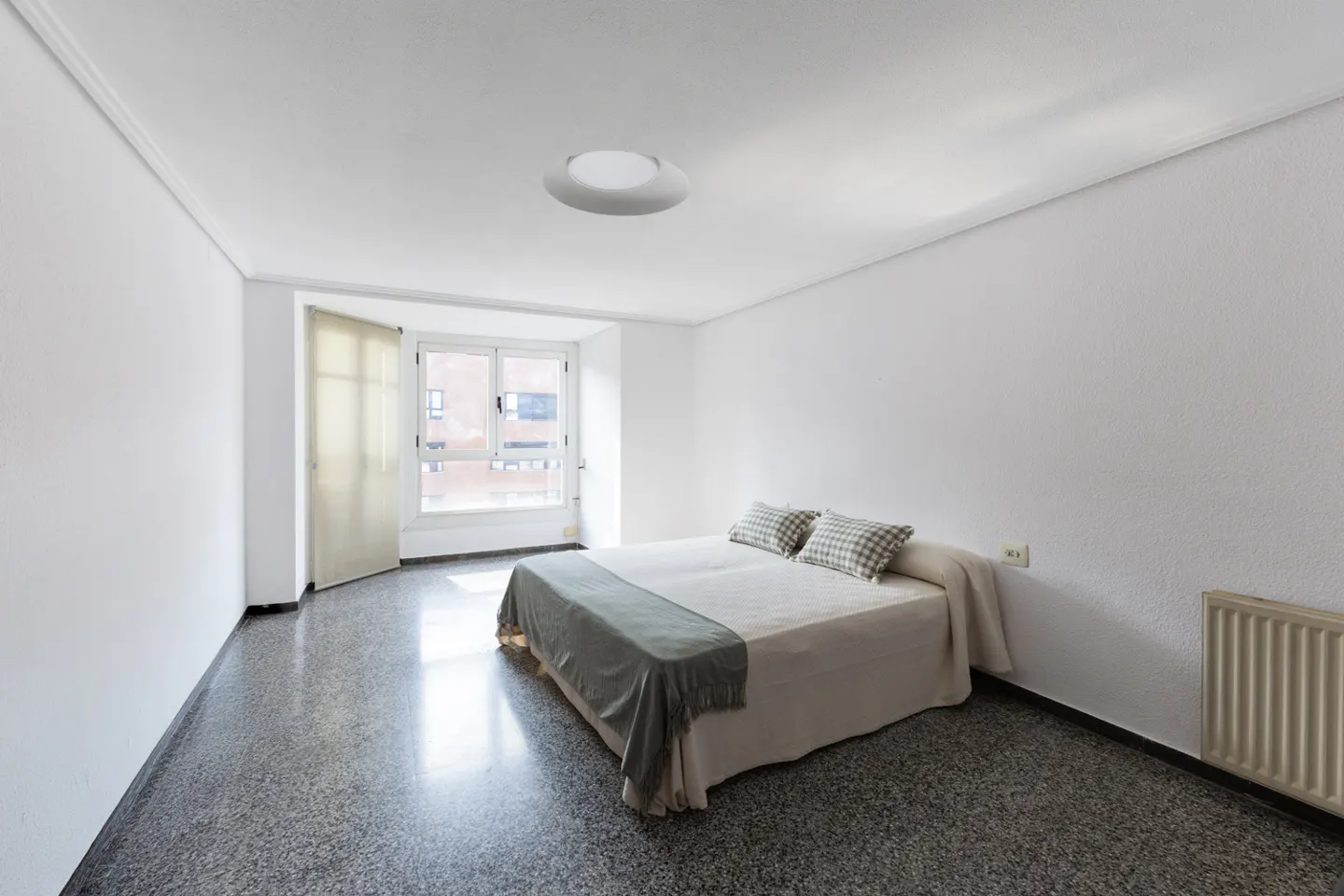 Bright bedroom with white walls, a double bed with gray accents, and a large window with sheer curtains. The floor is gray terrazzo.