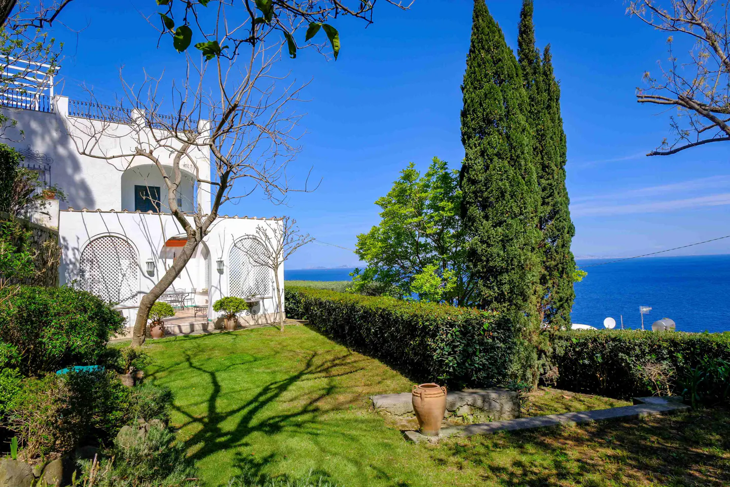 View of a white villa with arched windows, green lawn, trees, and the blue sea in the background.