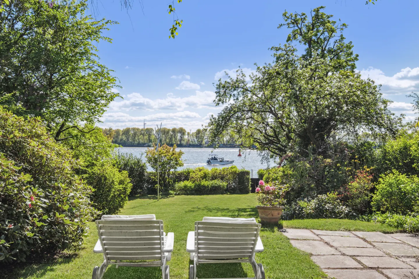 A backyard with two white lounge chairs facing a river with a boat on a sunny day.