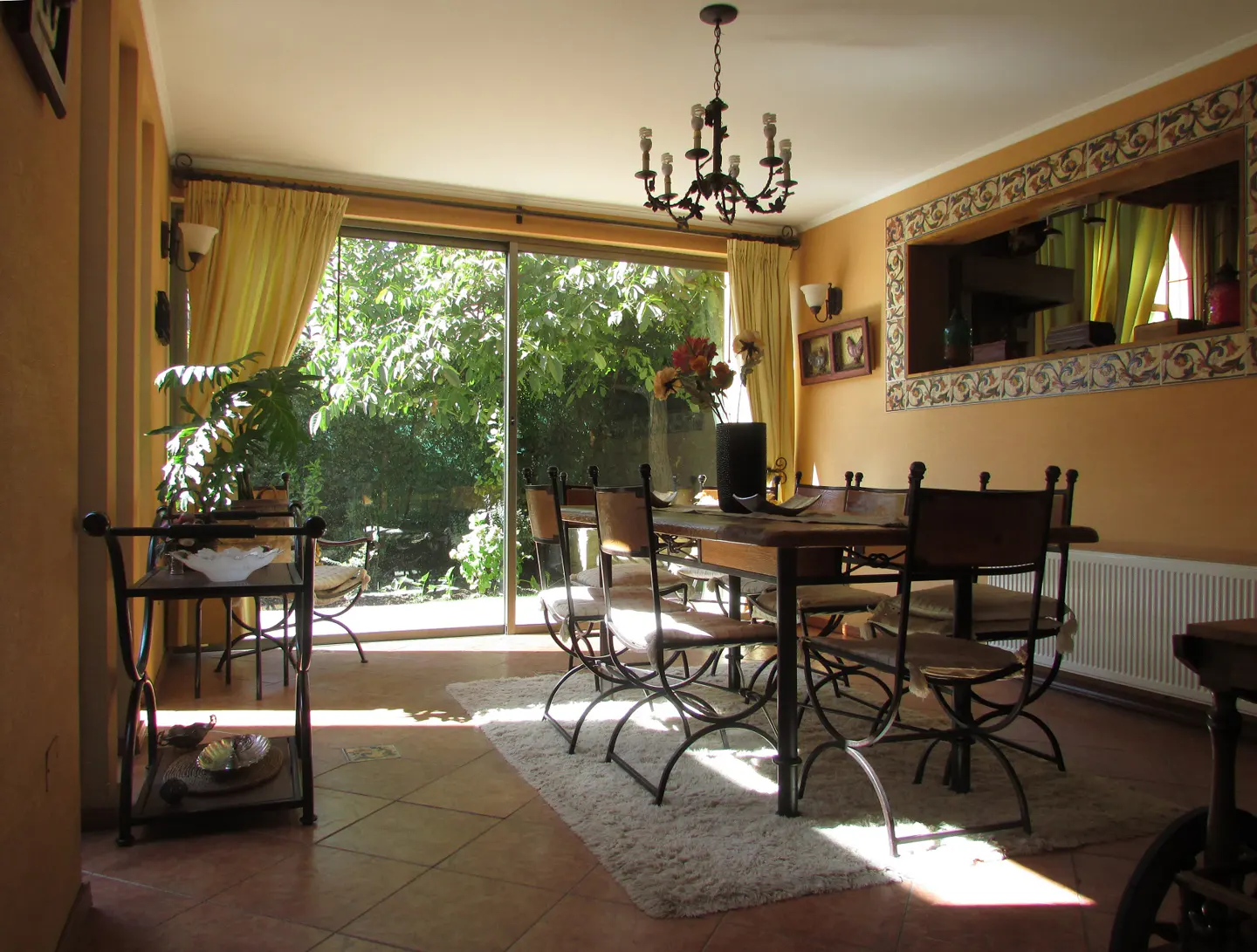 Dining room with a wooden table, chairs, and chandelier. Large windows overlook a lush green garden. The walls are peach-colored.