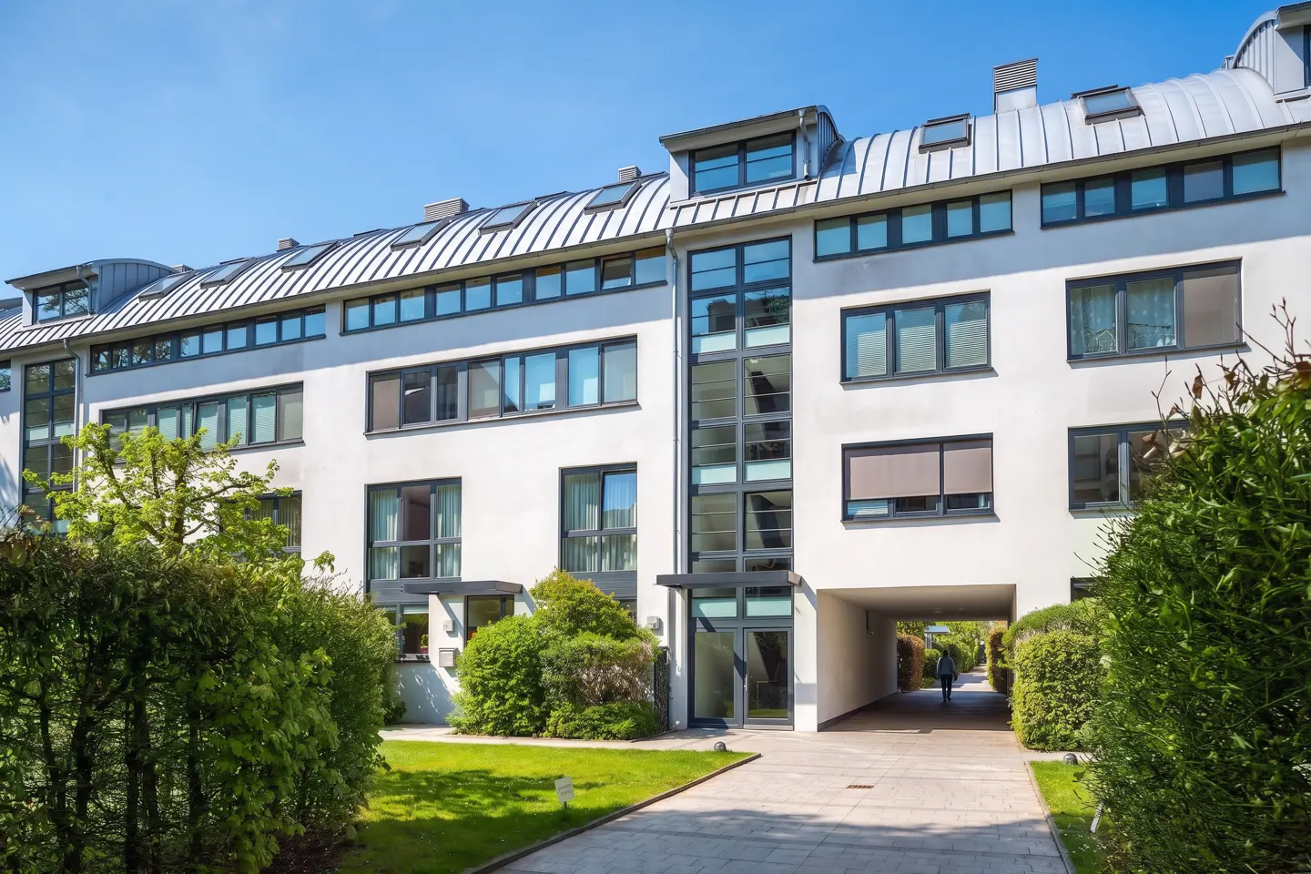 Modern white apartment building with a curved metal roof, large windows, and a paved walkway leading to a central entrance.