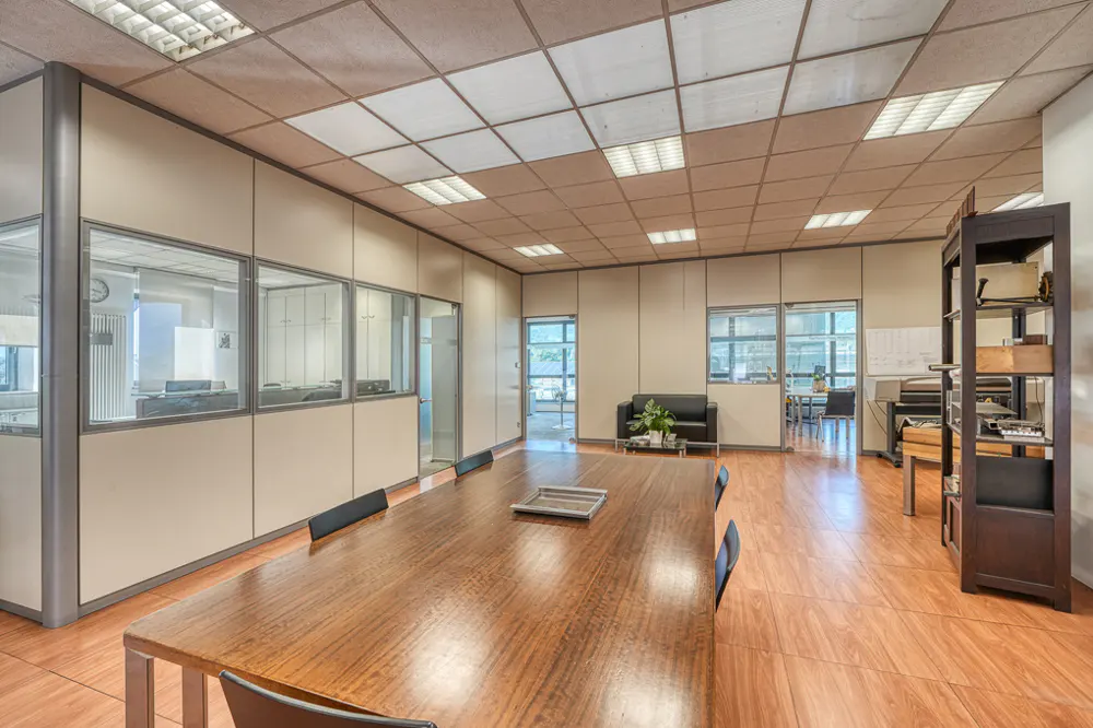 Office space with a long wooden conference table, glass-walled offices, and a black sofa in the background.