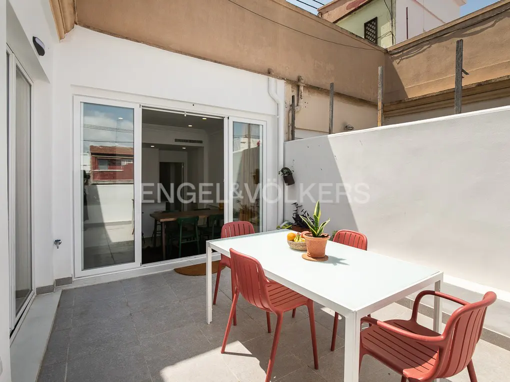 Outdoor patio with white table, four red chairs, and potted plant. Sliding glass doors lead to a dining room. Gray tile floor.