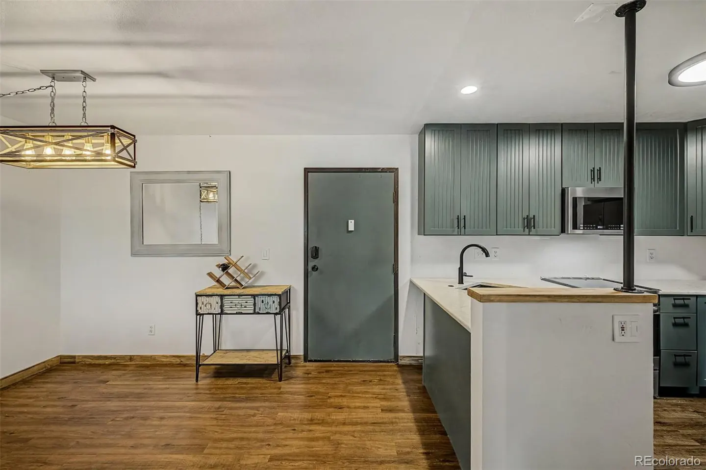 A modern kitchen with wood floors, white walls, and green cabinets. A gray door is next to a table with a wine rack.