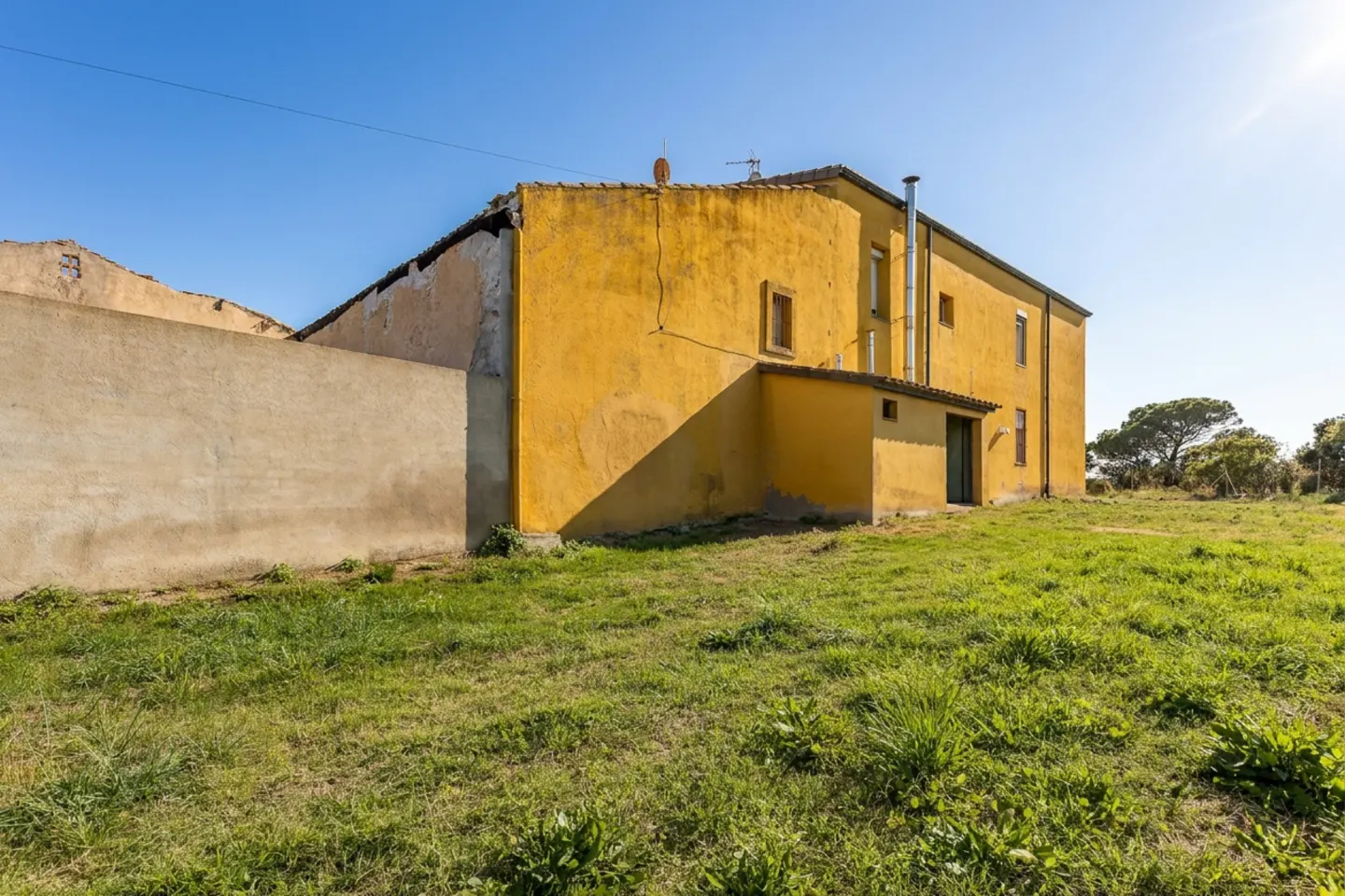 Exterior view of a two-story yellow house with a chimney, a concrete wall, and a green lawn under a clear blue sky.