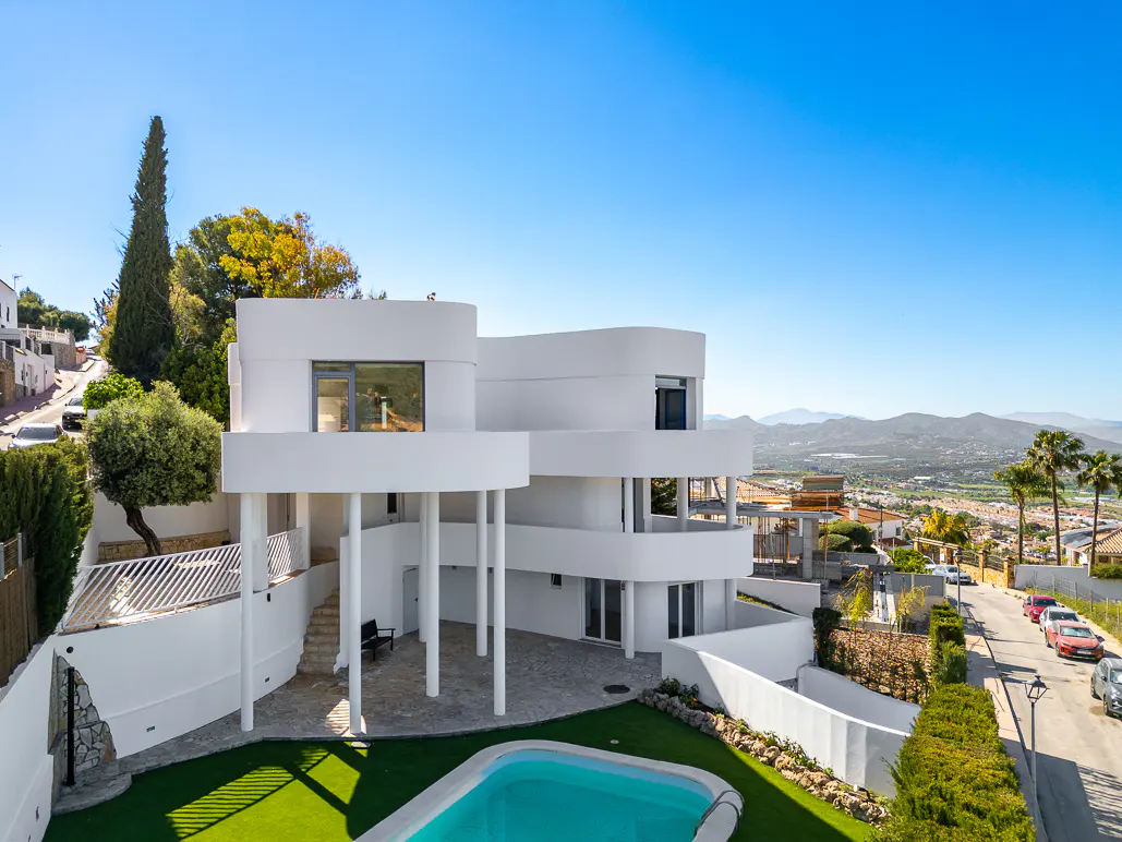 Modern white house with a pool, set against a clear blue sky and mountain backdrop. Lush greenery surrounds the property.