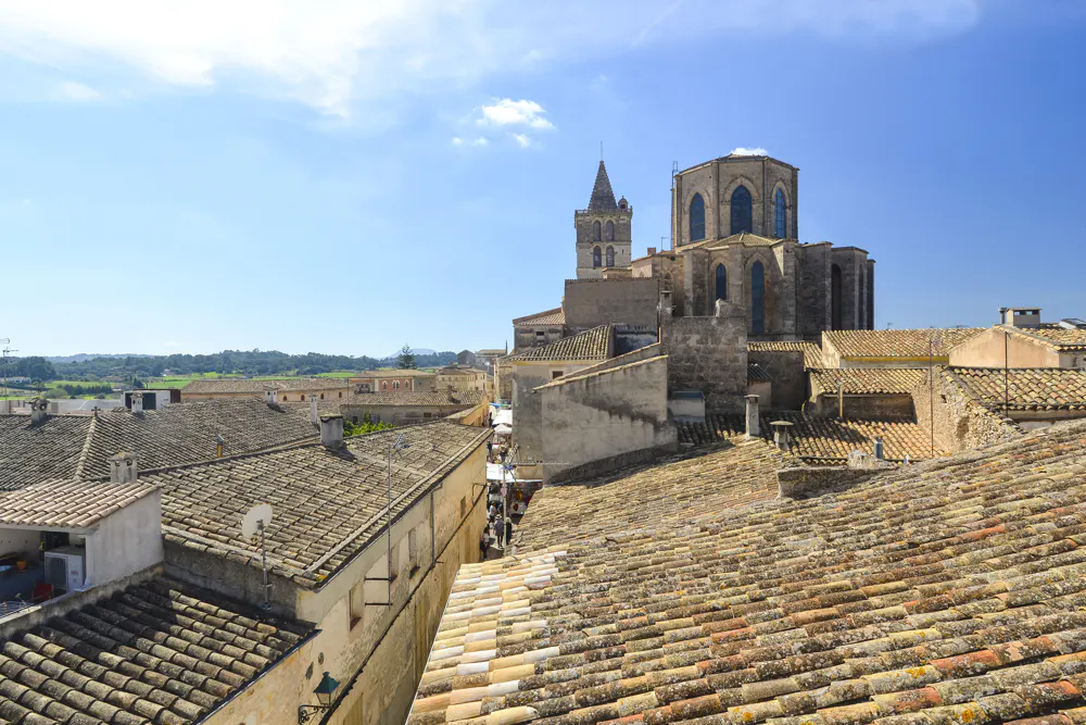 View of terracotta rooftops and a stone church with a tall spire under a blue sky in Mallorca, Spain.
