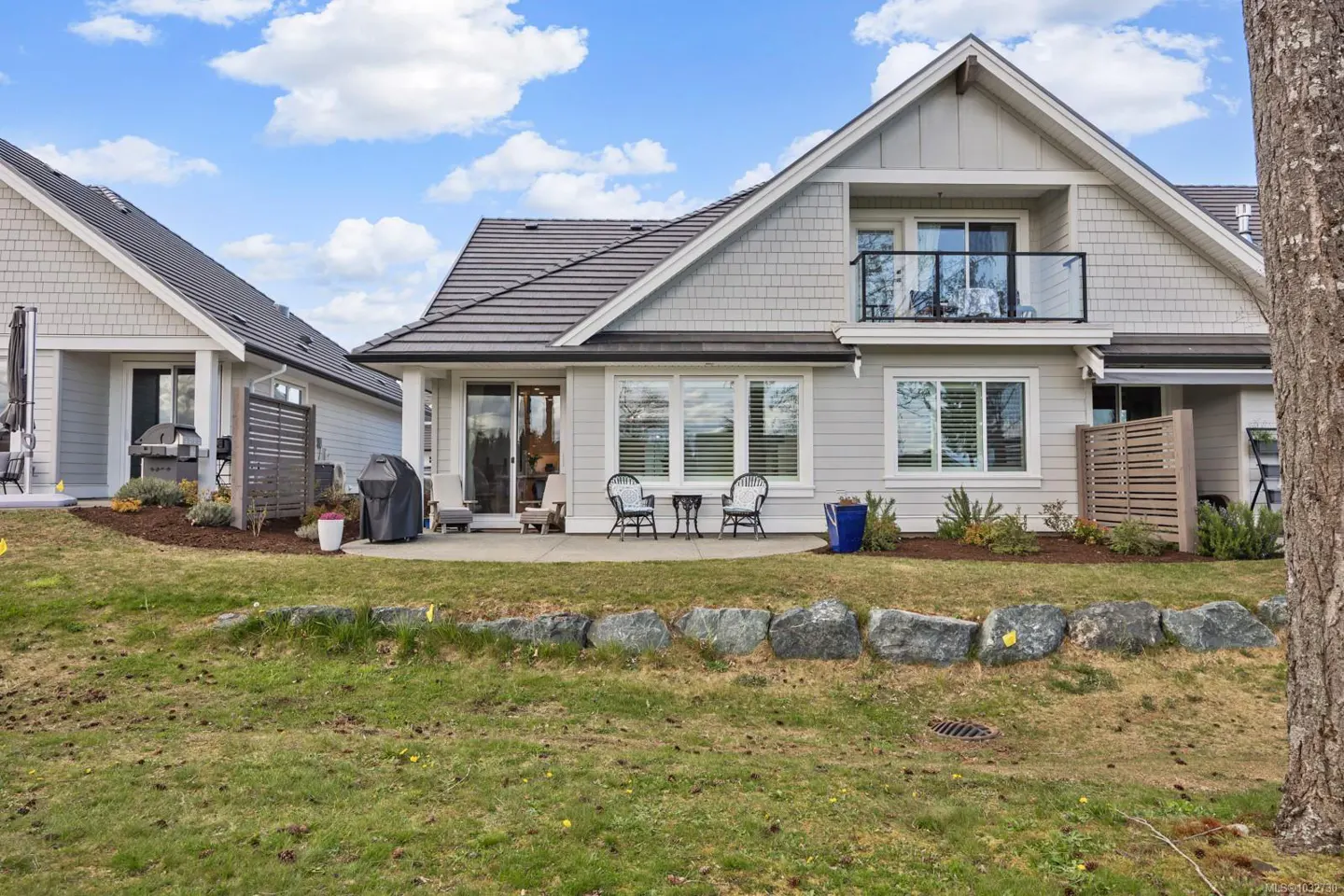 Exterior view of a two-story, light gray house with a balcony, patio, and green lawn.