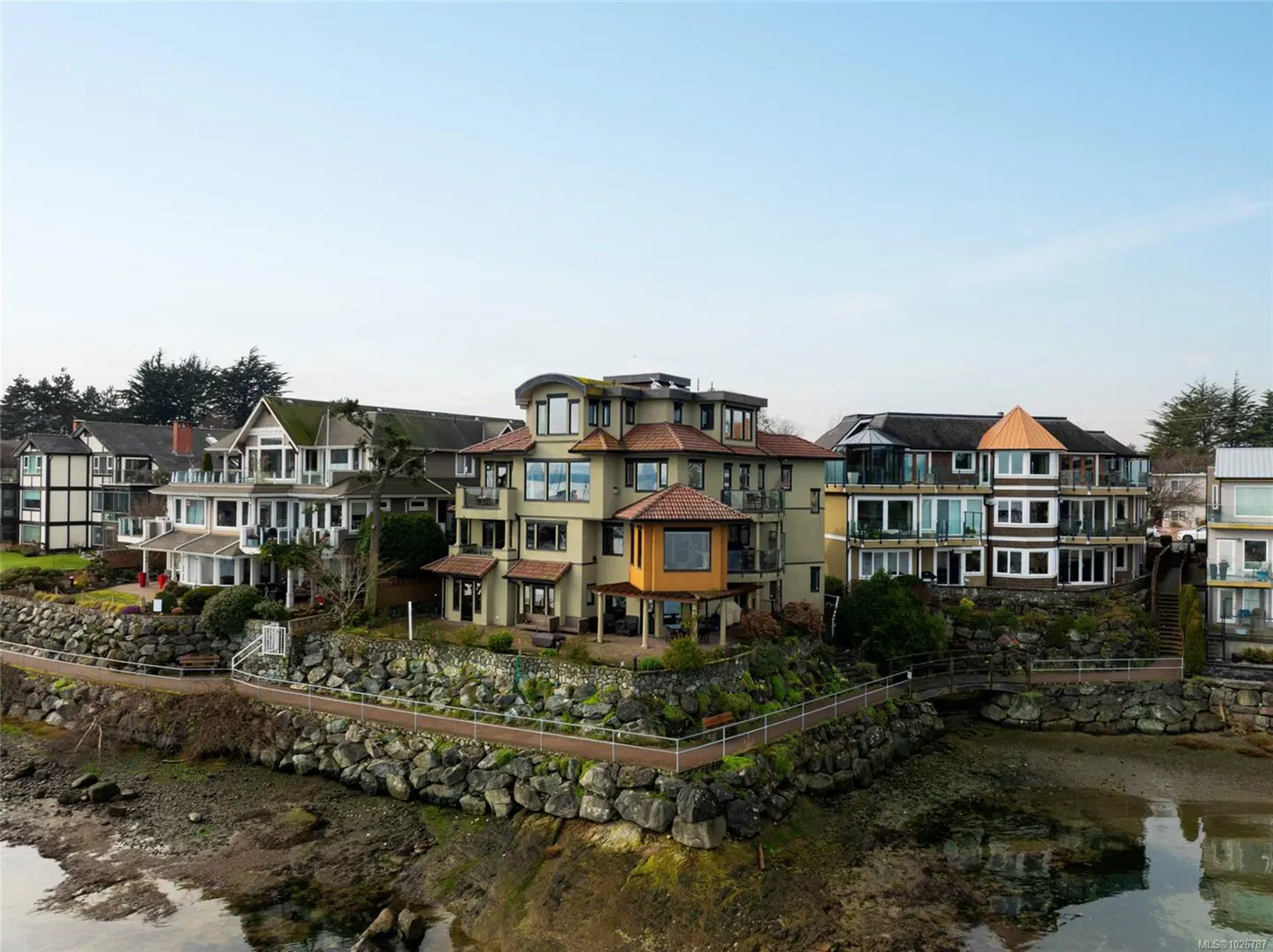 Waterfront homes with stone walls and a walkway line the coast under a clear sky.