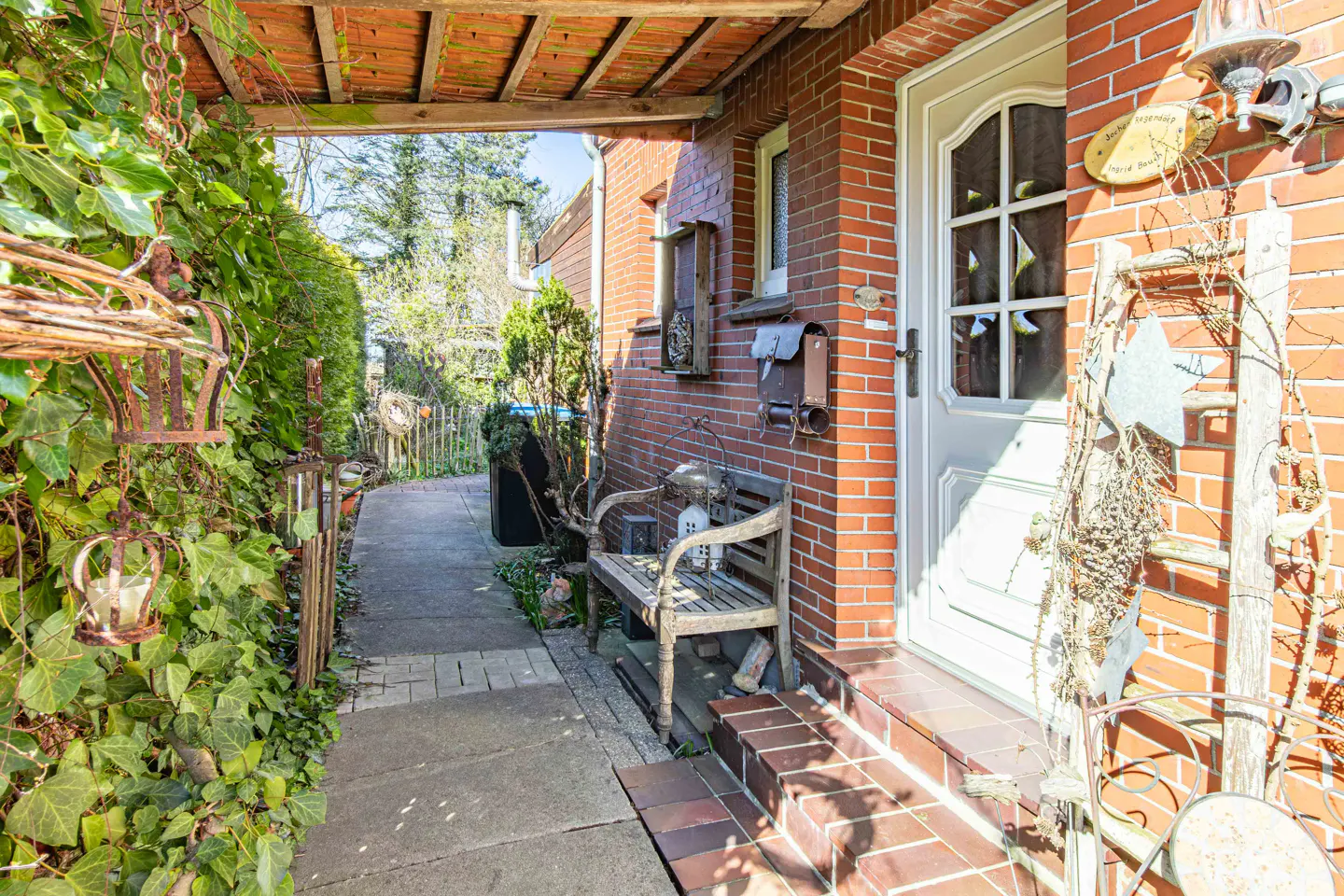 Brick house entrance with white door, bench, and ivy-covered walkway. A mailbox and rustic decor add charm.