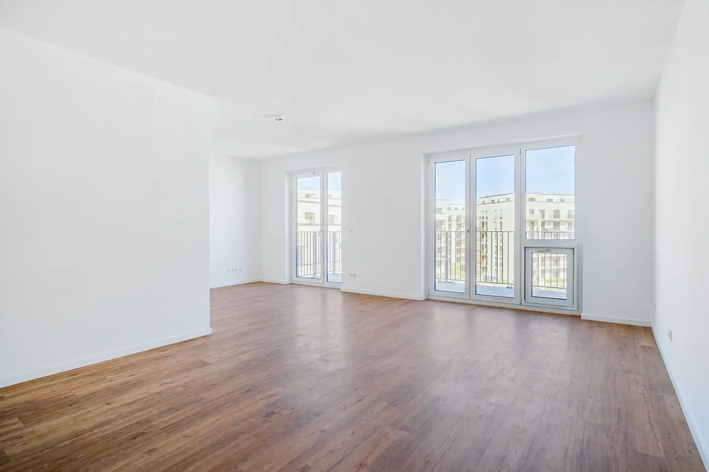 Bright, empty room with wood floors, white walls, and two sets of glass doors leading to balconies.