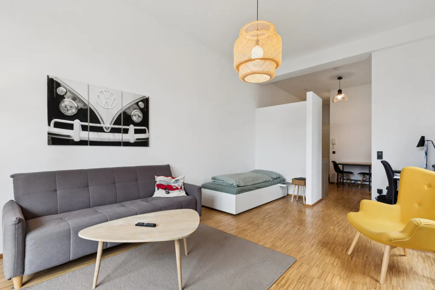 Living room with gray sofa, wood floor, and yellow chair. A VW bus print hangs above the sofa. A bed is in the background.