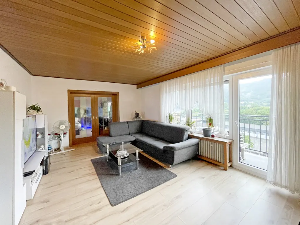 Living room with gray sectional sofa, glass coffee table on a gray rug. Wood paneled ceiling, white walls, and balcony with sheer curtains.
